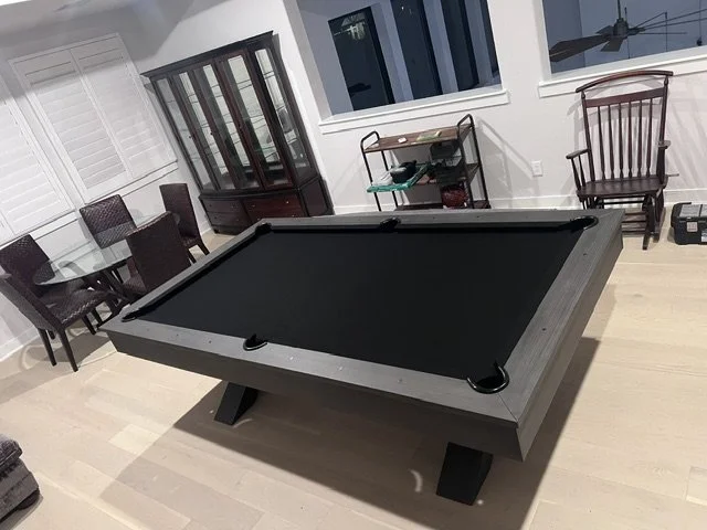 A black pool table in a dining room with chairs, a wooden display cabinet, and a window.