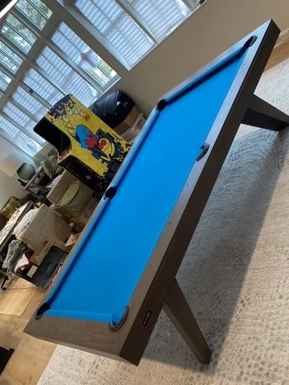 Indoor scene with a blue pool table, a yellow armchair with colorful patterns, and shelves with various items in the background.