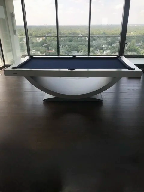 Indoor room with a modern pool table near large floor-to-ceiling windows revealing a cityscape view.