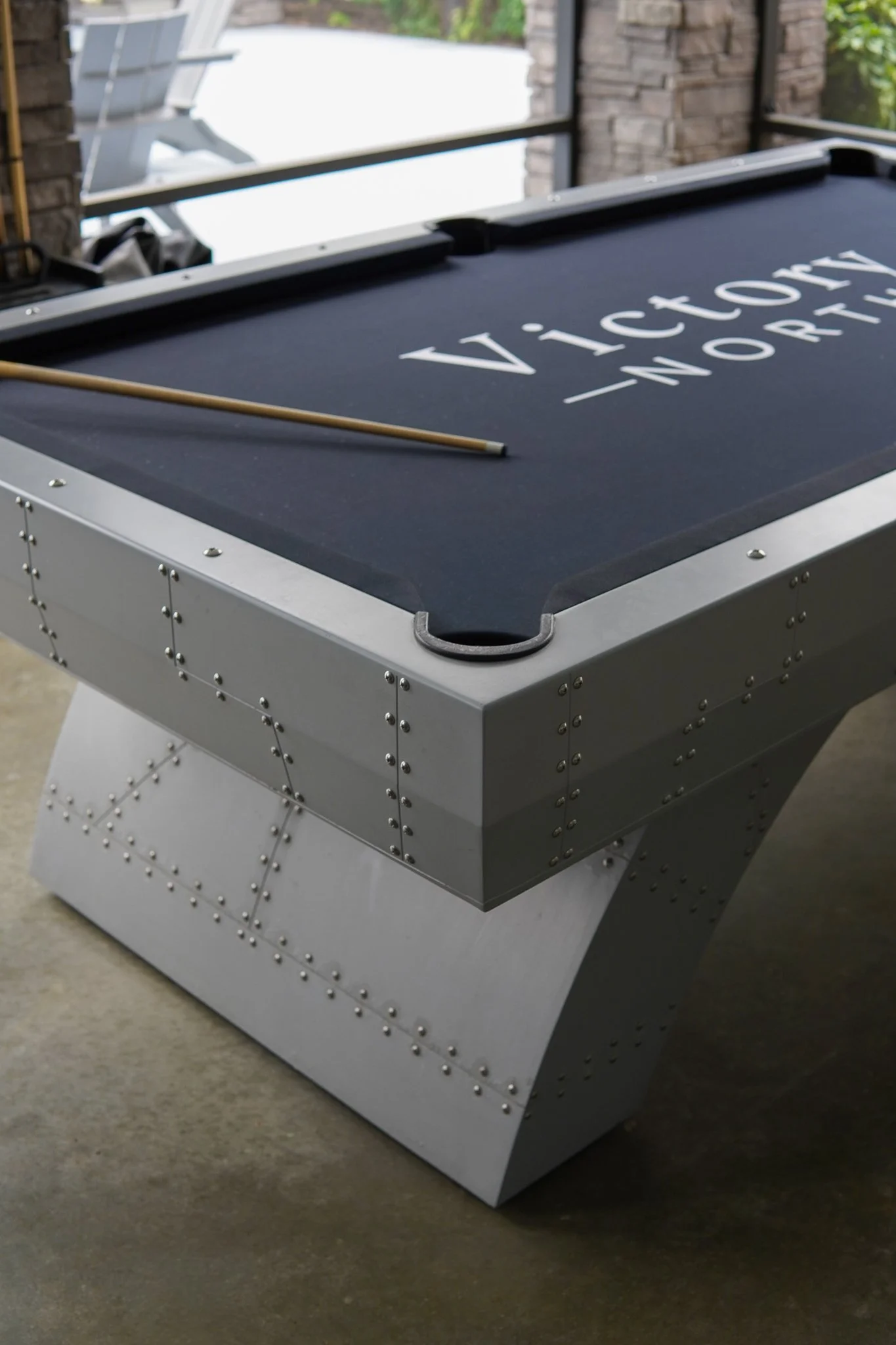 An air hockey table with a black playing surface and a gold cue stick resting on it, located indoors near brick walls and large windows.