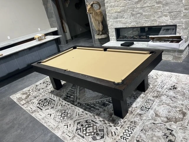A room with a pool table with beige felt, black frame, and four legs, placed on a patterned area rug. In the background, there is a stone fireplace with a glass front, a deer head mounted on the wall, and a white shelf with boxes.