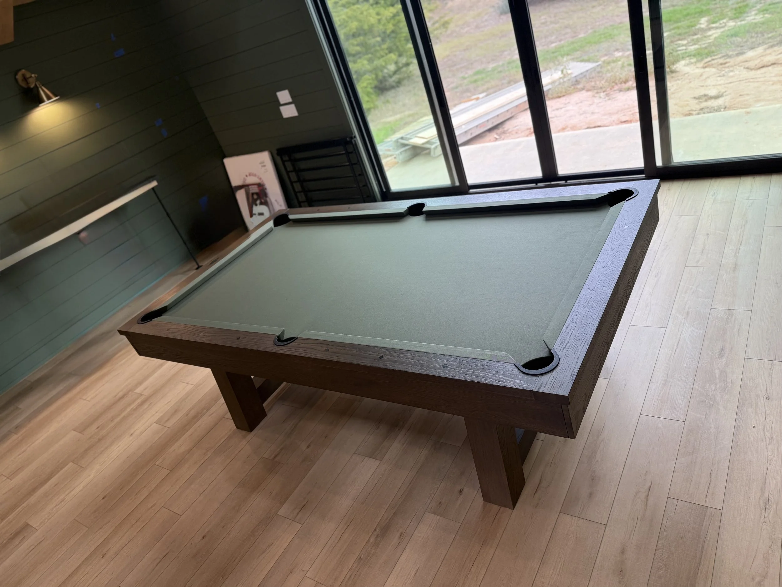 A pool table with green felt and wooden frame inside a room with wood flooring, near large glass sliding doors that open to an outdoor area with dirt and grass.