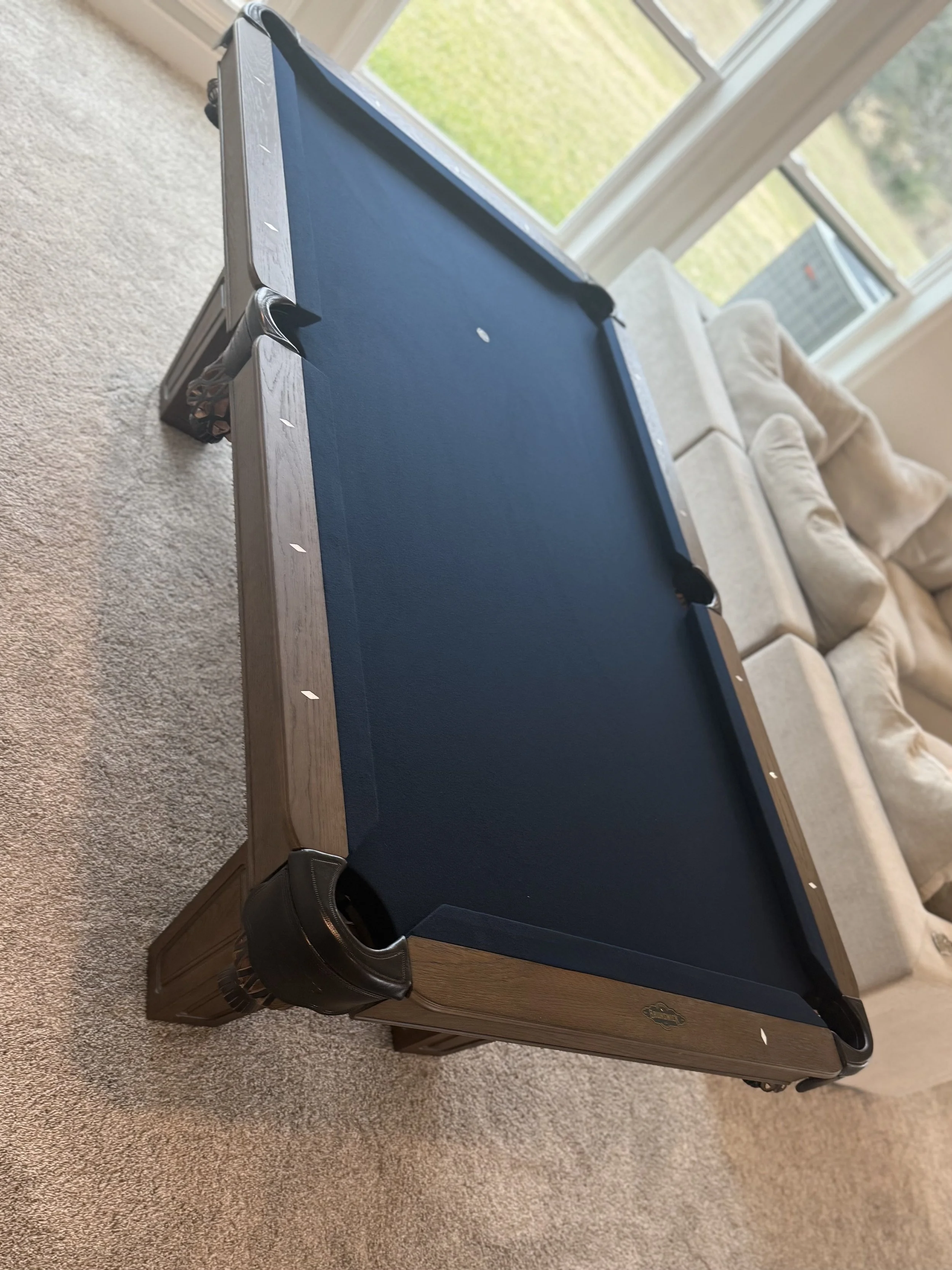 A pool table with a blue felt surface in a room with large windows and a beige sofa in the background.