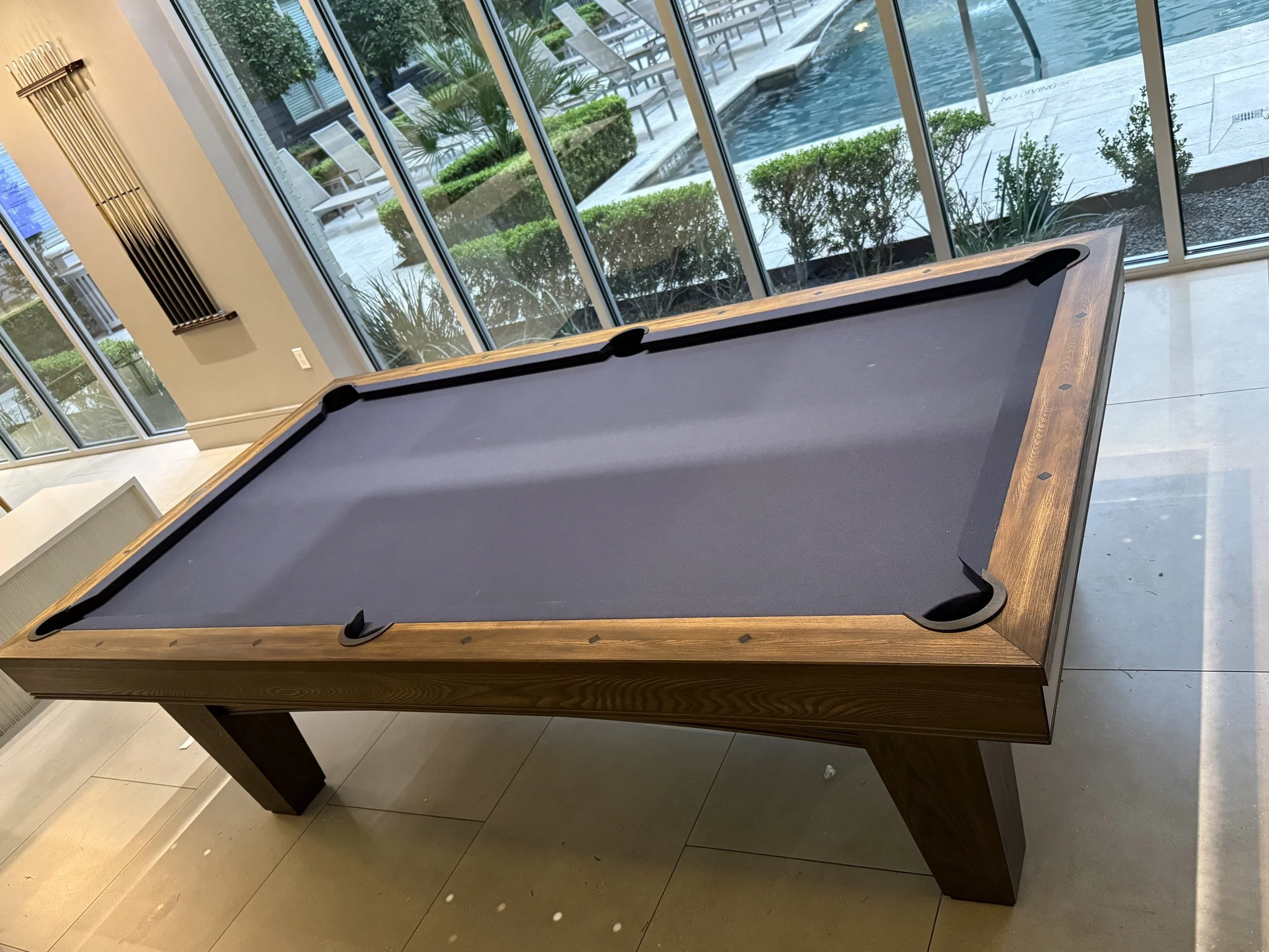 Indoor pool table with padded playing surface in a room with large glass windows overlooking an outdoor pool and lounge chairs.