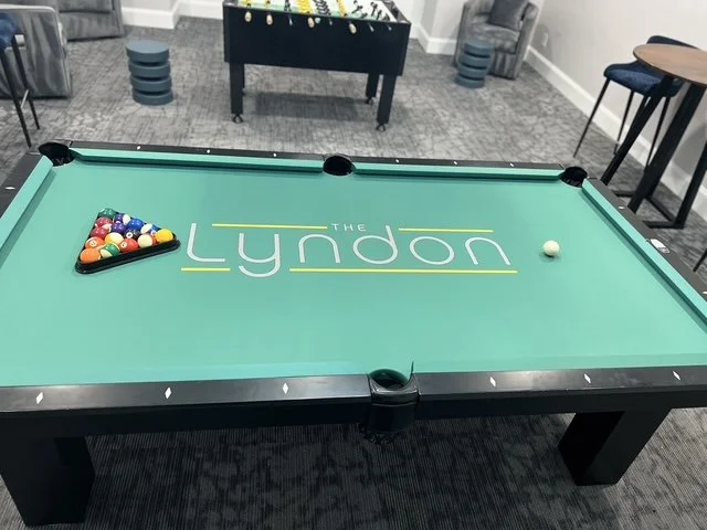 A pool table with a triangle of pool balls and a cue ball on it, in a recreational room with chairs and a foosball table in the background.