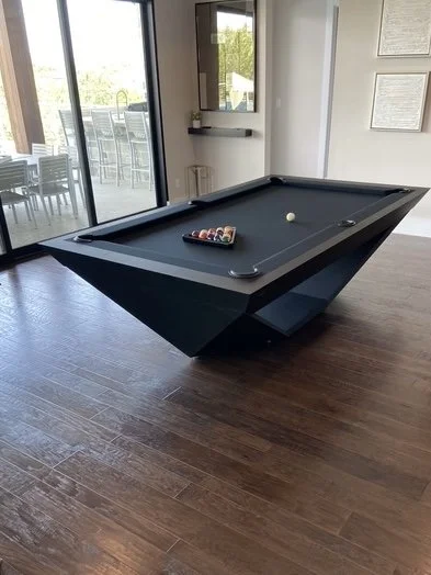 A black pool table with balls and cue ball on a wooden floor next to a glass sliding door.