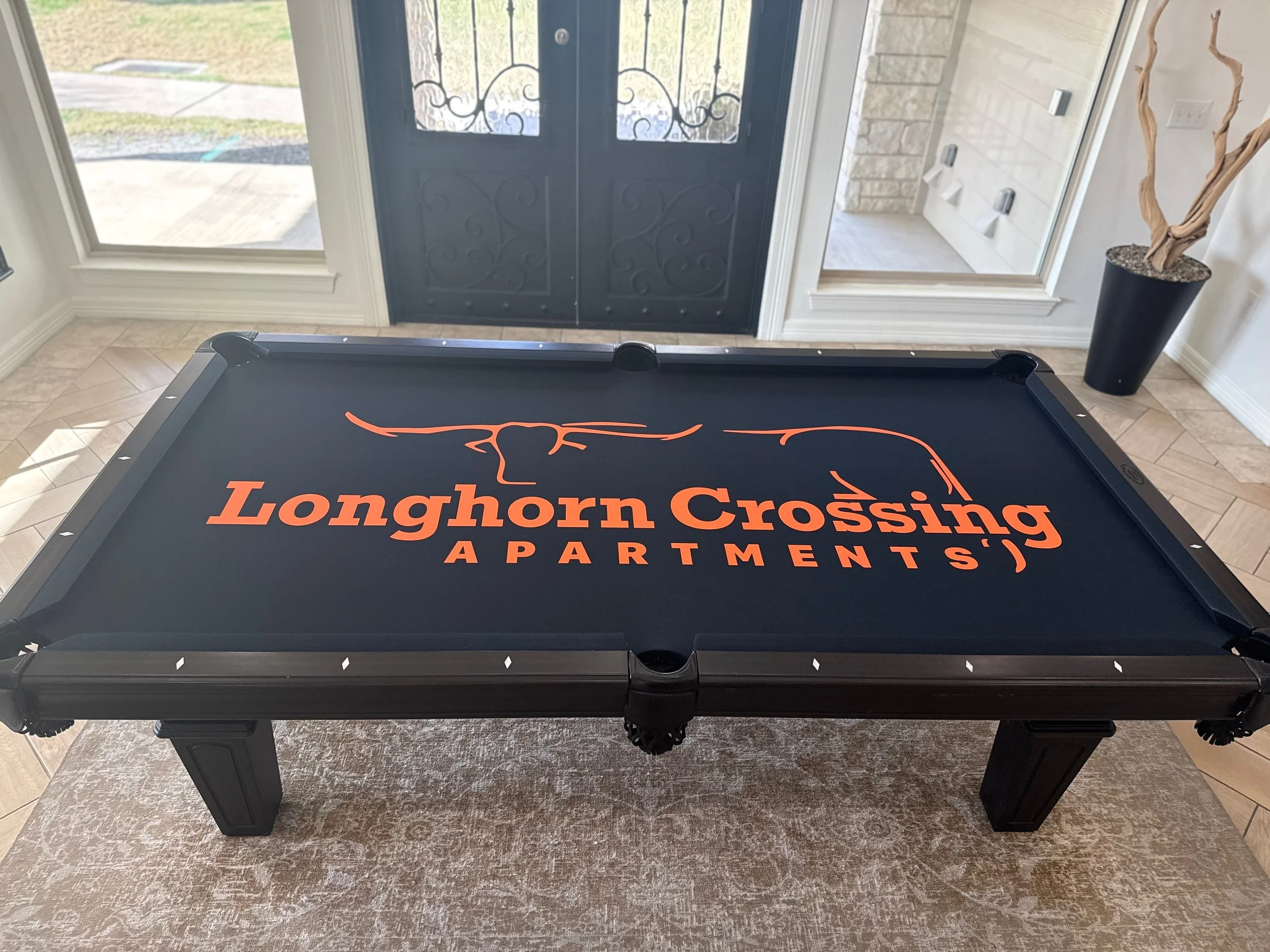 Indoor view of an entrance with two black decorative front doors with wrought iron designs, side windows, a corner with a tall black planter containing a twisty dried branch, and a pool table with a Longhorn Crossfire logo.