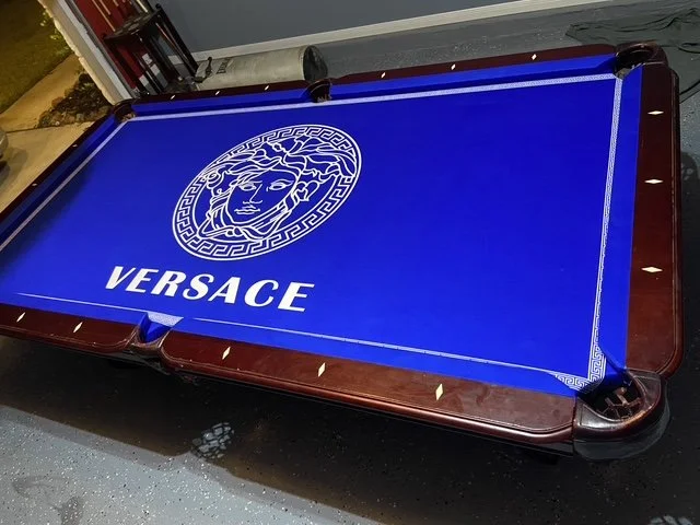 A pool table with a bright blue felt playing surface featuring the Versace logo and branding.