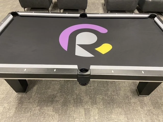 An overhead view of a black pool table with a purple, white, and yellow abstract logo in the center, and black chairs in the background.