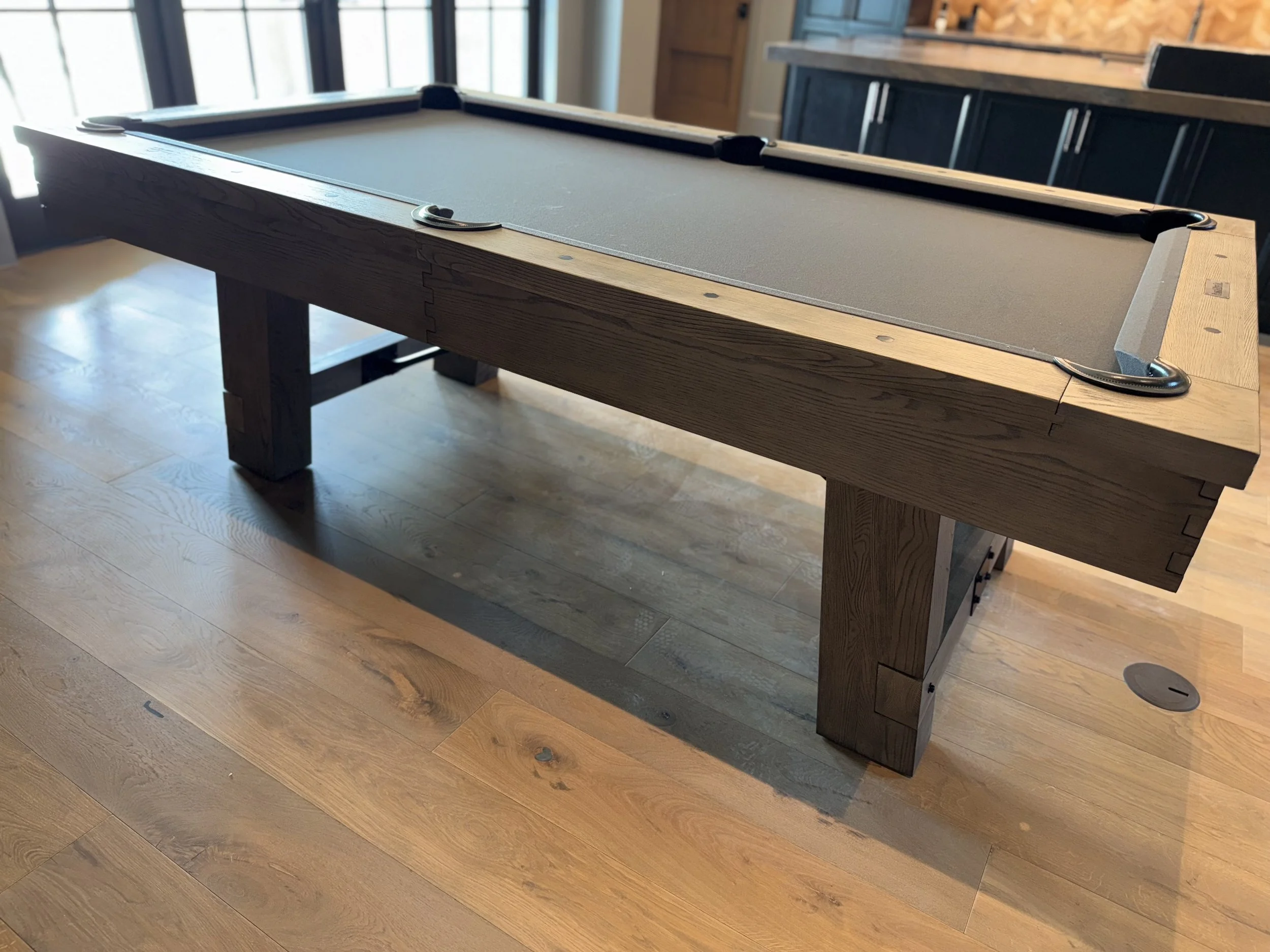 A pool table with gray felt and wooden frame in a room with hardwood floors and natural light from large windows.