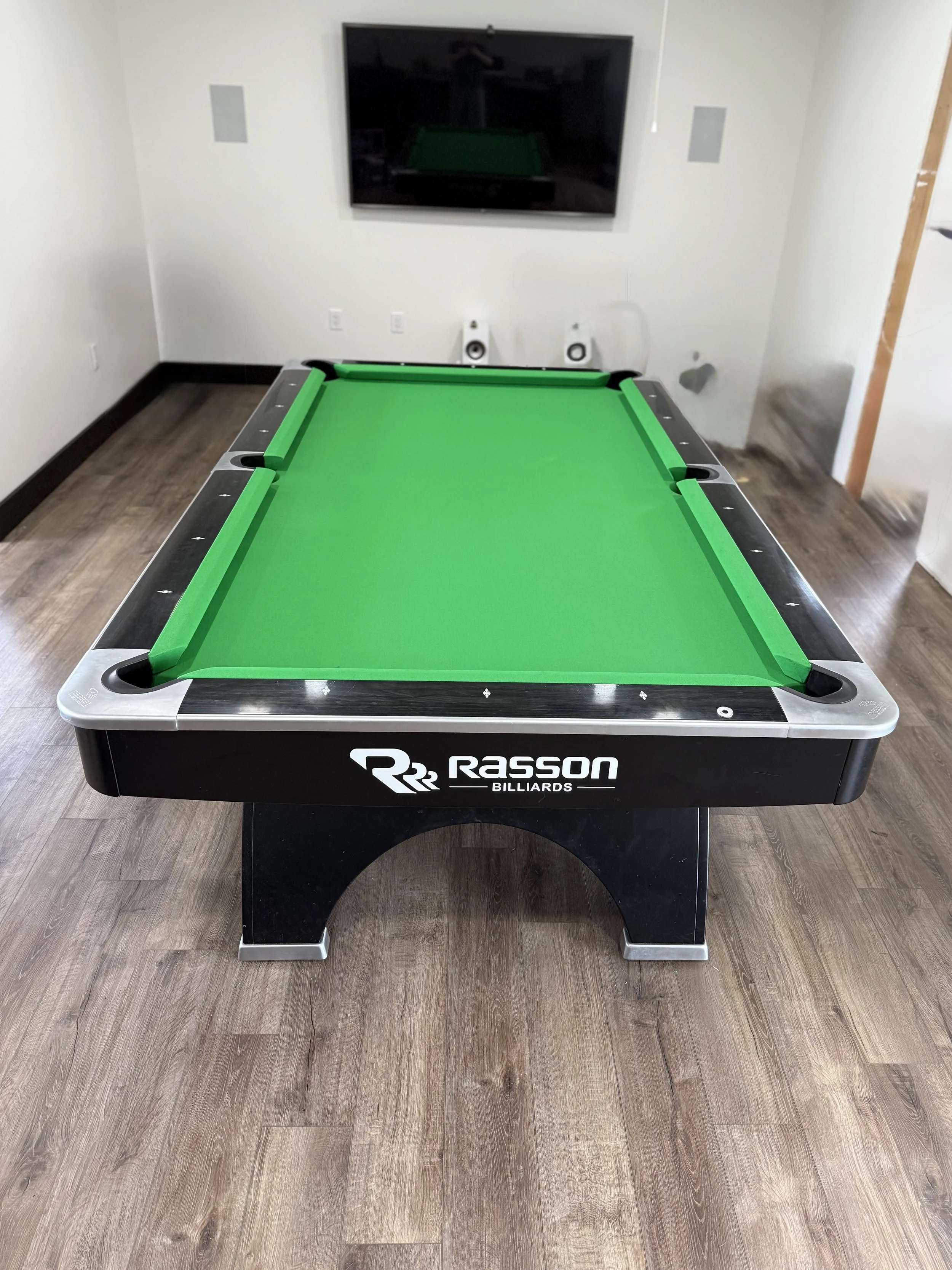 An indoor air hockey table with a green playing surface and black sides, positioned on a wooden floor in front of a wall.