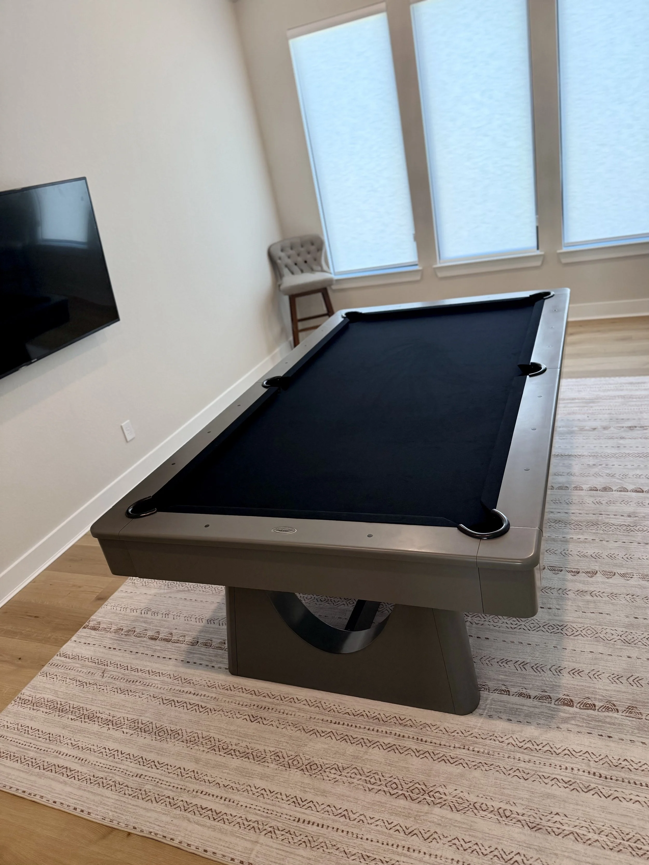 A bedroom with a pool table on a patterned beige and white rug, a wall-mounted TV, a beige tufted chair near large windows with light blue blinds.