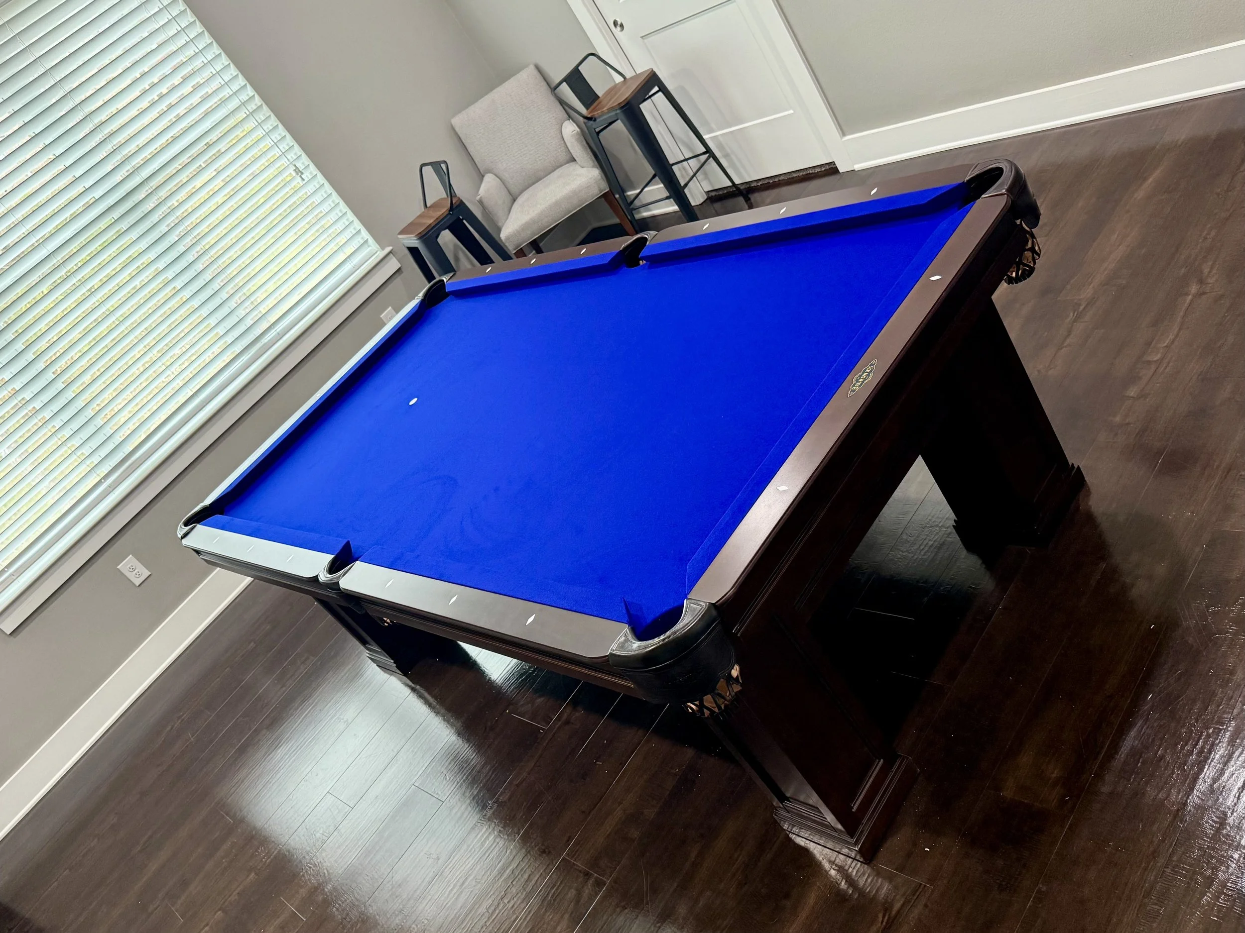 A room with a pool table with a blue felt top, a beige armchair, a small black and wood side table, and a window with closed white blinds. The floor is dark hardwood.