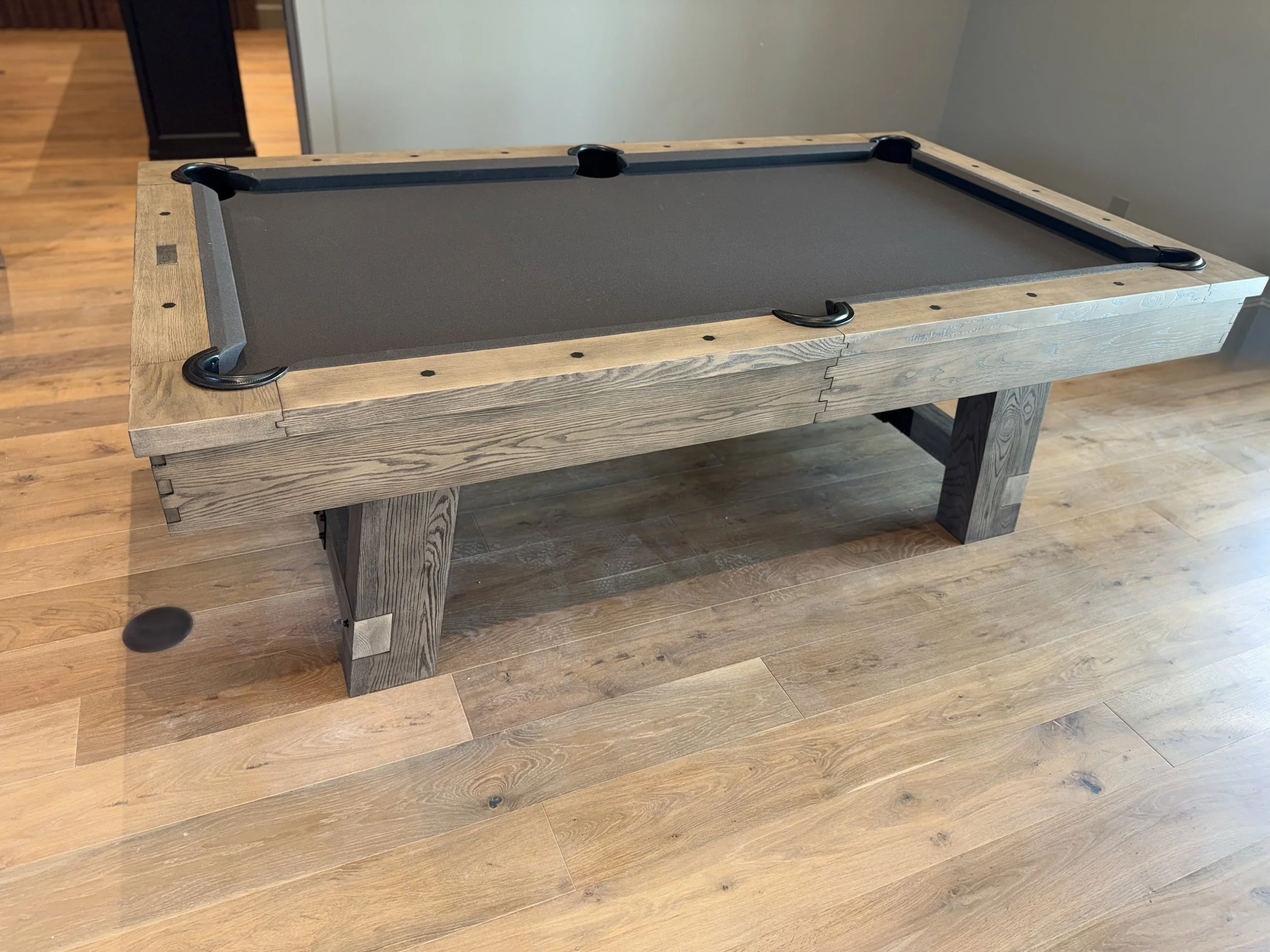 A pool table with a wooden frame and black felt surface, placed in a room with wooden flooring and plain light-colored walls.