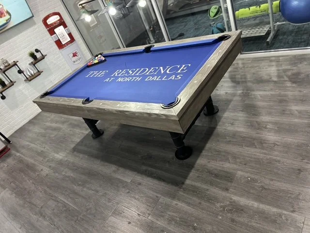 Pool table with blue felt and the words 'The Residence at North Dallas' printed on it, located in a recreational room.