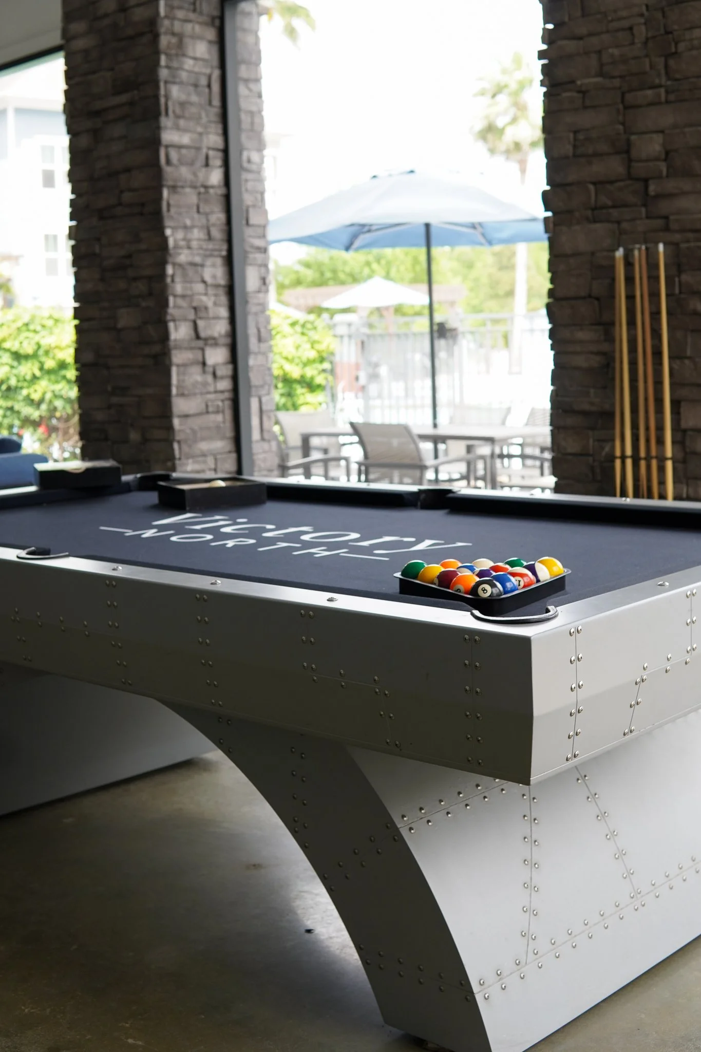 A pool table with pool balls arranged in a triangle, situated near large windows with stone pillars and an outdoor patio with umbrellas.