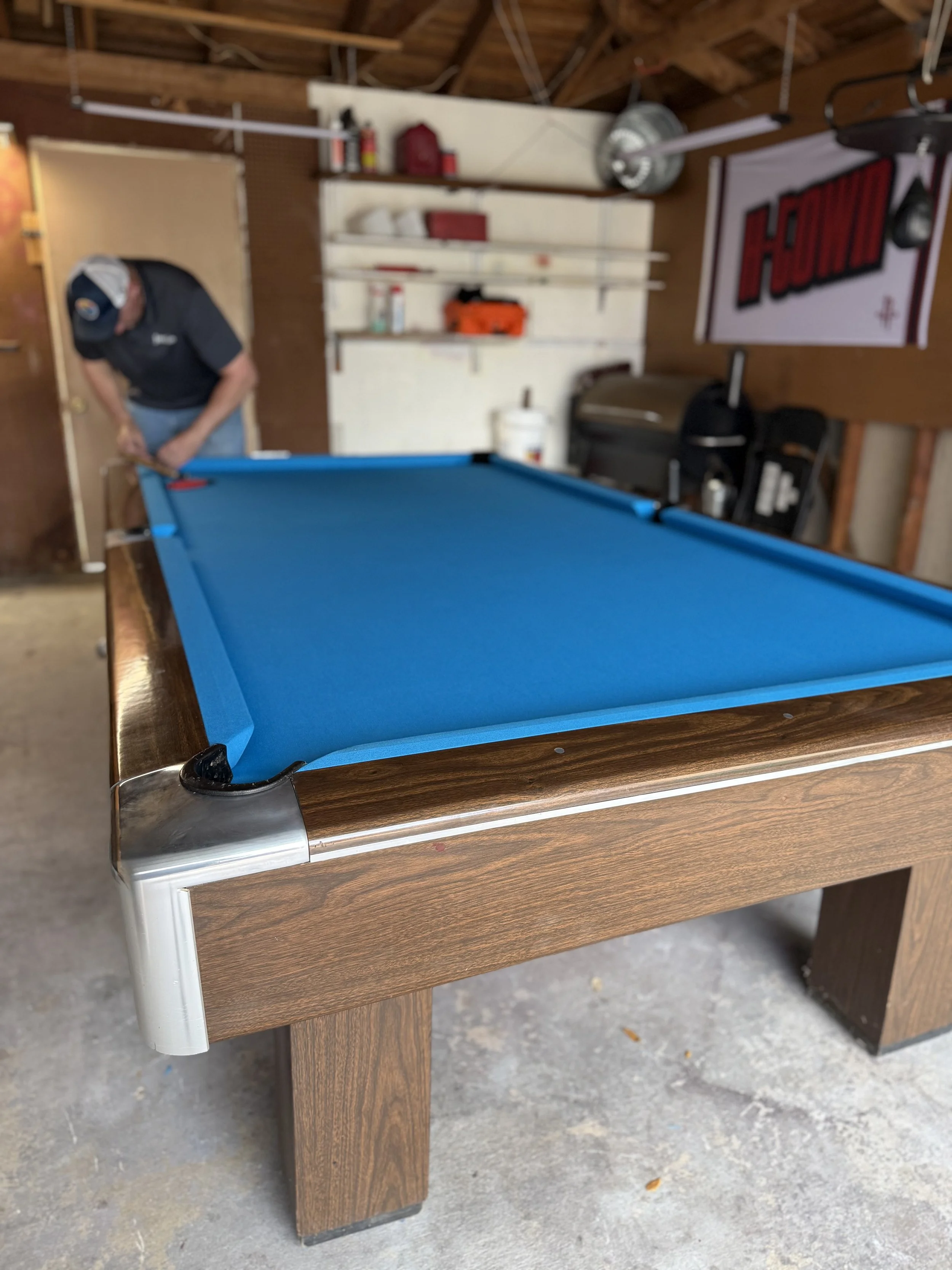 A pool table with a blue felt surface in a garage or basement setting, showing a man in a black cap and shirt standing near the table, with tools and shelves on the wall behind him and a partially visible stool or chair beside the table.