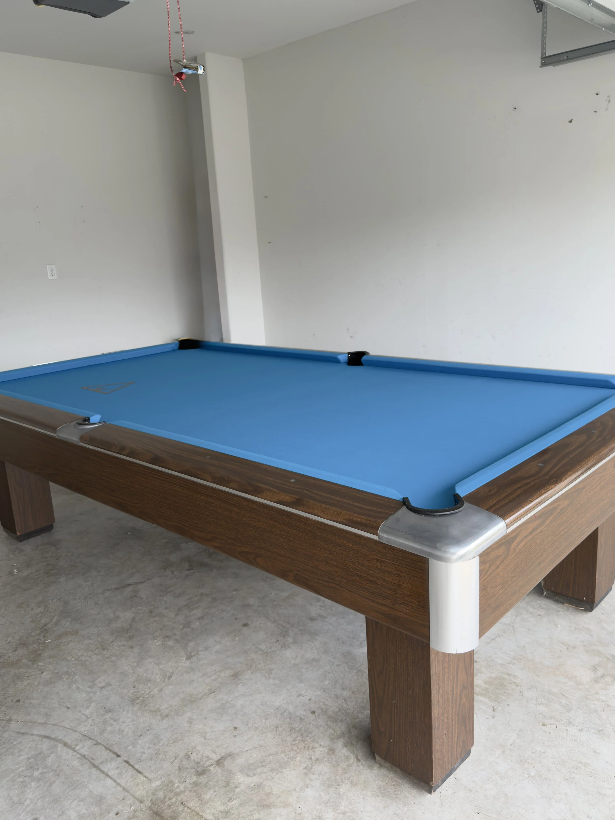 A pool table with a blue felt surface and wooden sides in a garage with plain white walls and a concrete floor.
