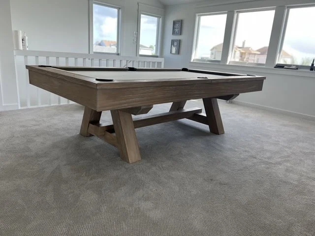 A wooden pool table in a bright, empty room with large windows and beige carpet.
