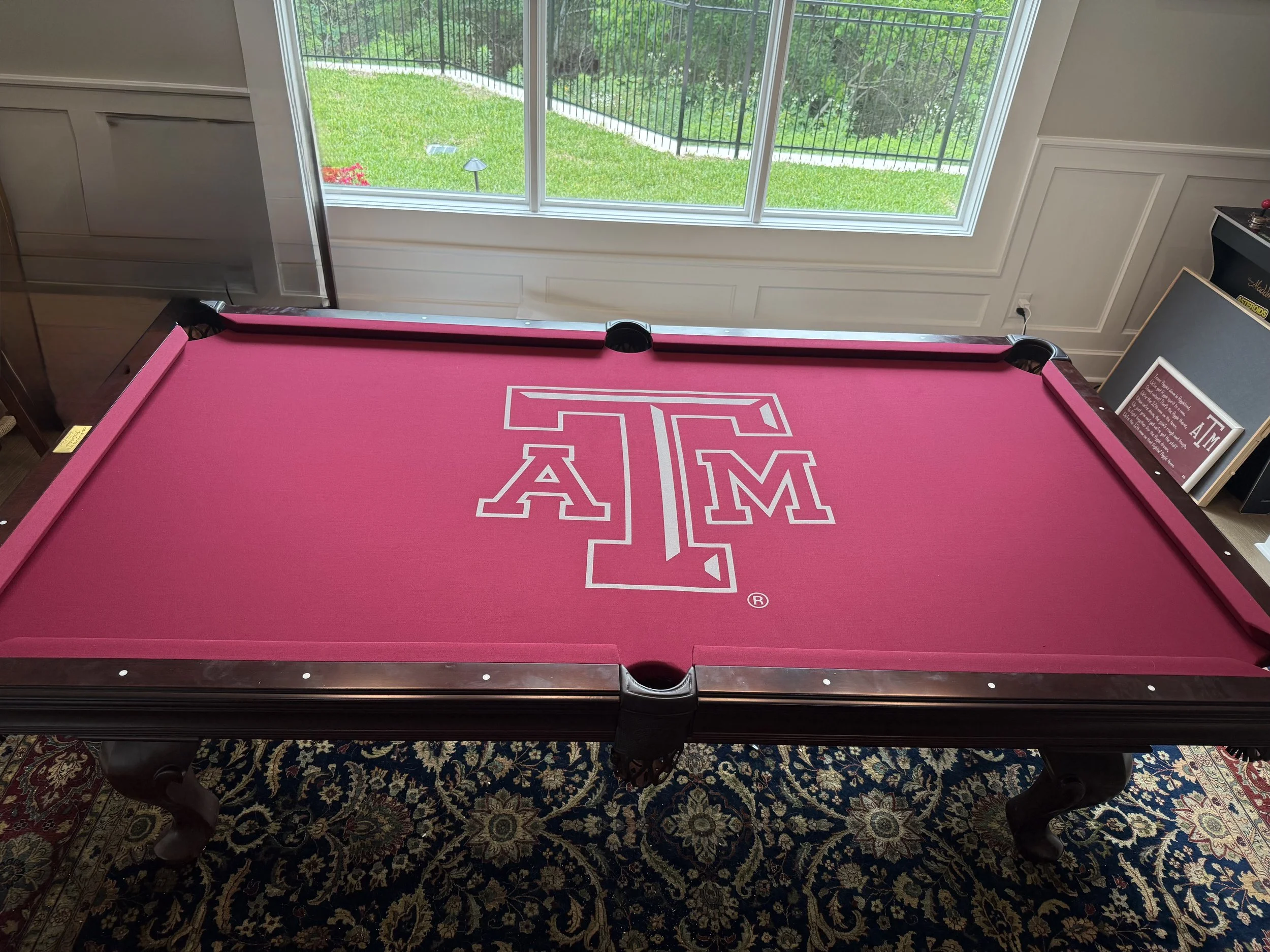 A pool table with a pink felt surface and a white A and M logo at the center. The table is indoors, near a large window looking out to a lush green backyard with a fence and trees.