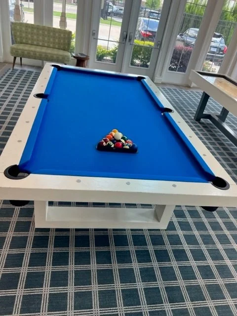 Indoor pool table with a blue felt surface and a triangle rack of billiard balls near the center, in a room with large windows, patterned carpet, and a green armchair.