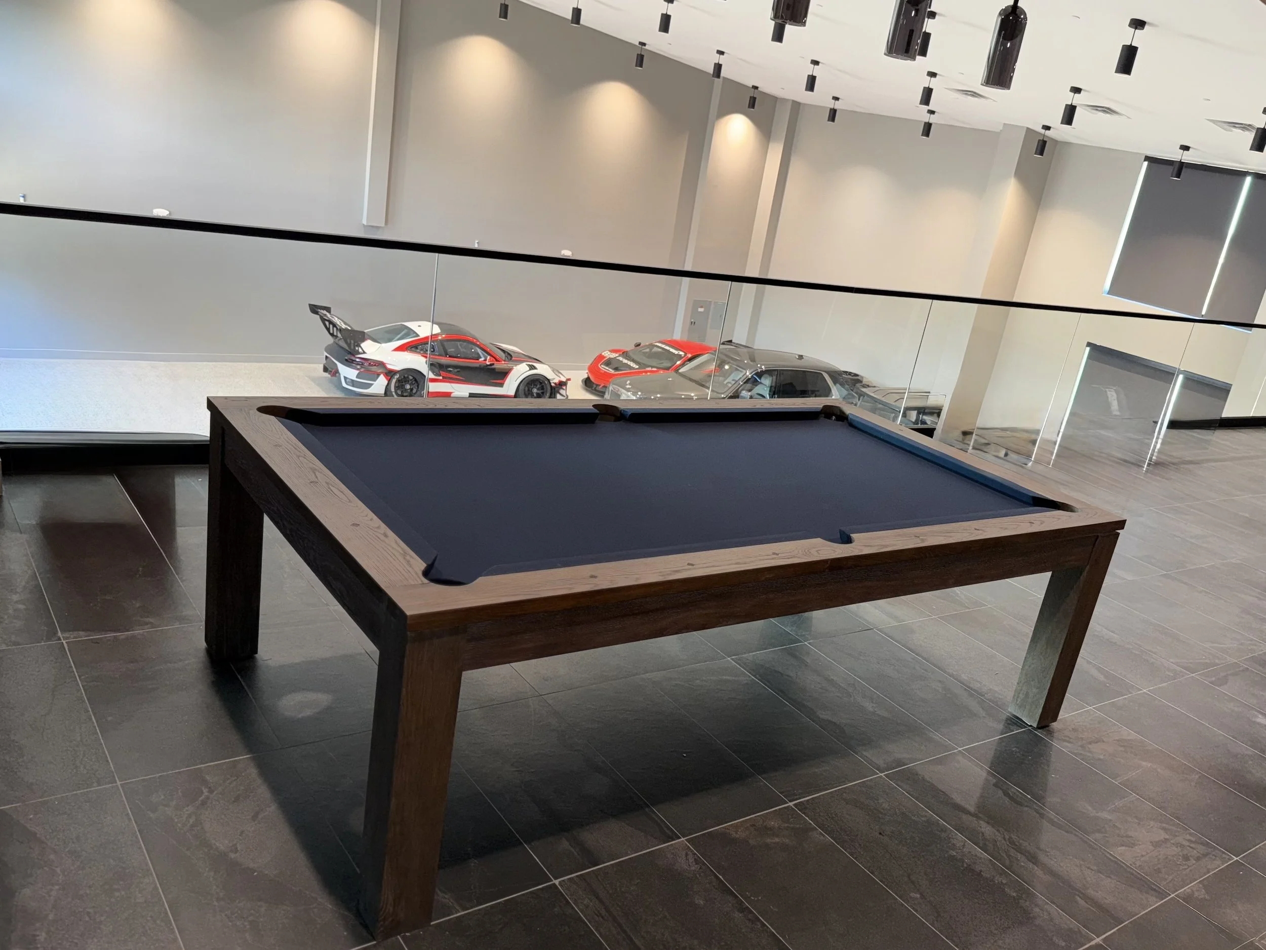 An indoor sports or recreational space featuring a pool table in the foreground and three high-end sports cars parked on a lowered level behind a glass barrier. The room has gray walls, black hanging light fixtures, and a modern, sleek design.