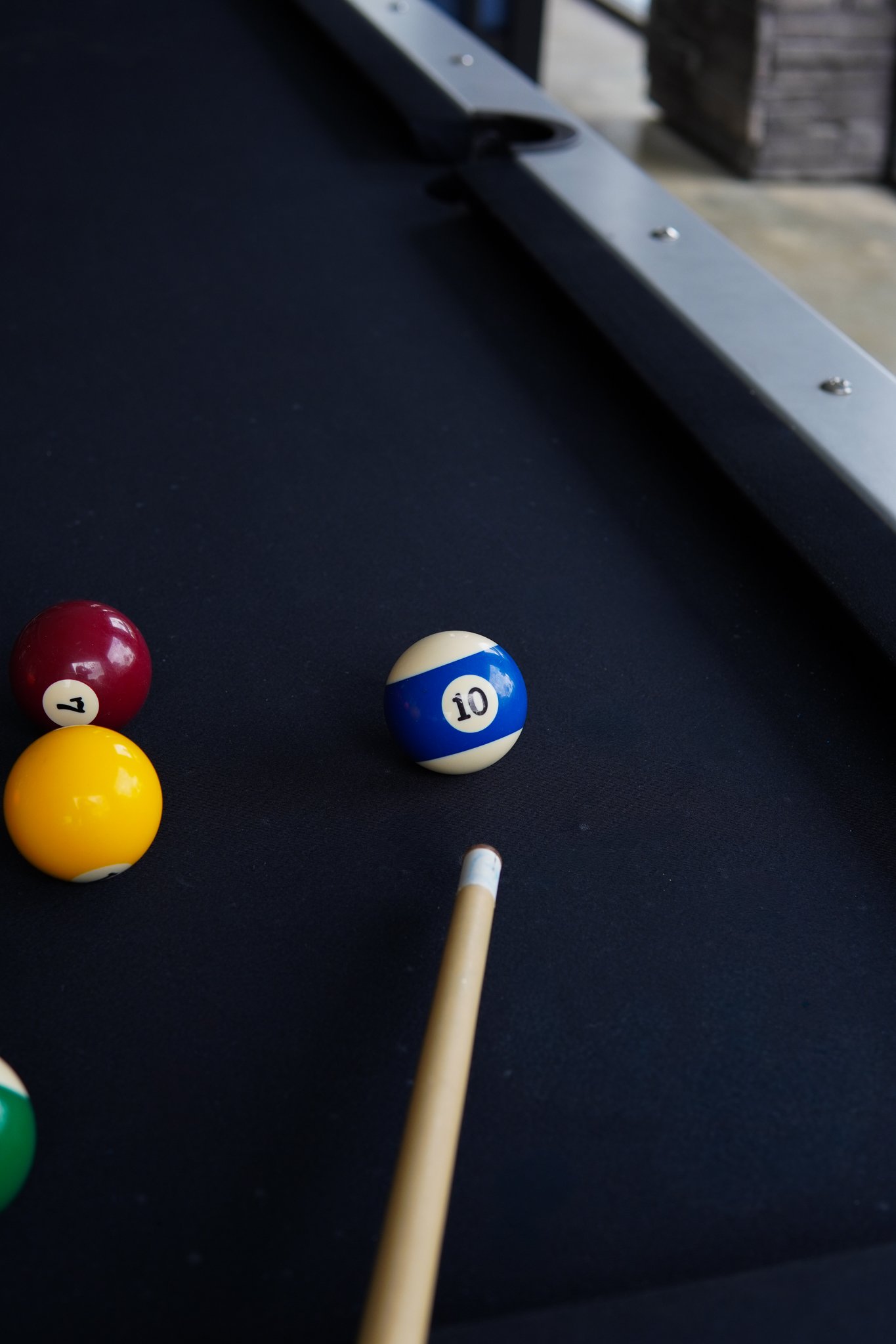 Close-up of pool balls on a black pool table, with only the 10 ball visible near the cue stick, and part of a metal cue rack in the background.