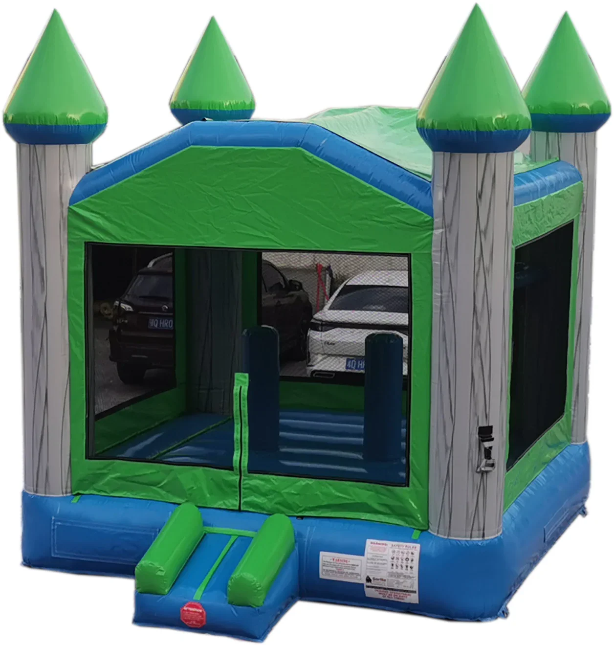 Colorful inflatable bounce house with castle design, featuring four green turrets and mesh windows, set up in a parking lot with cars visible through the windows.