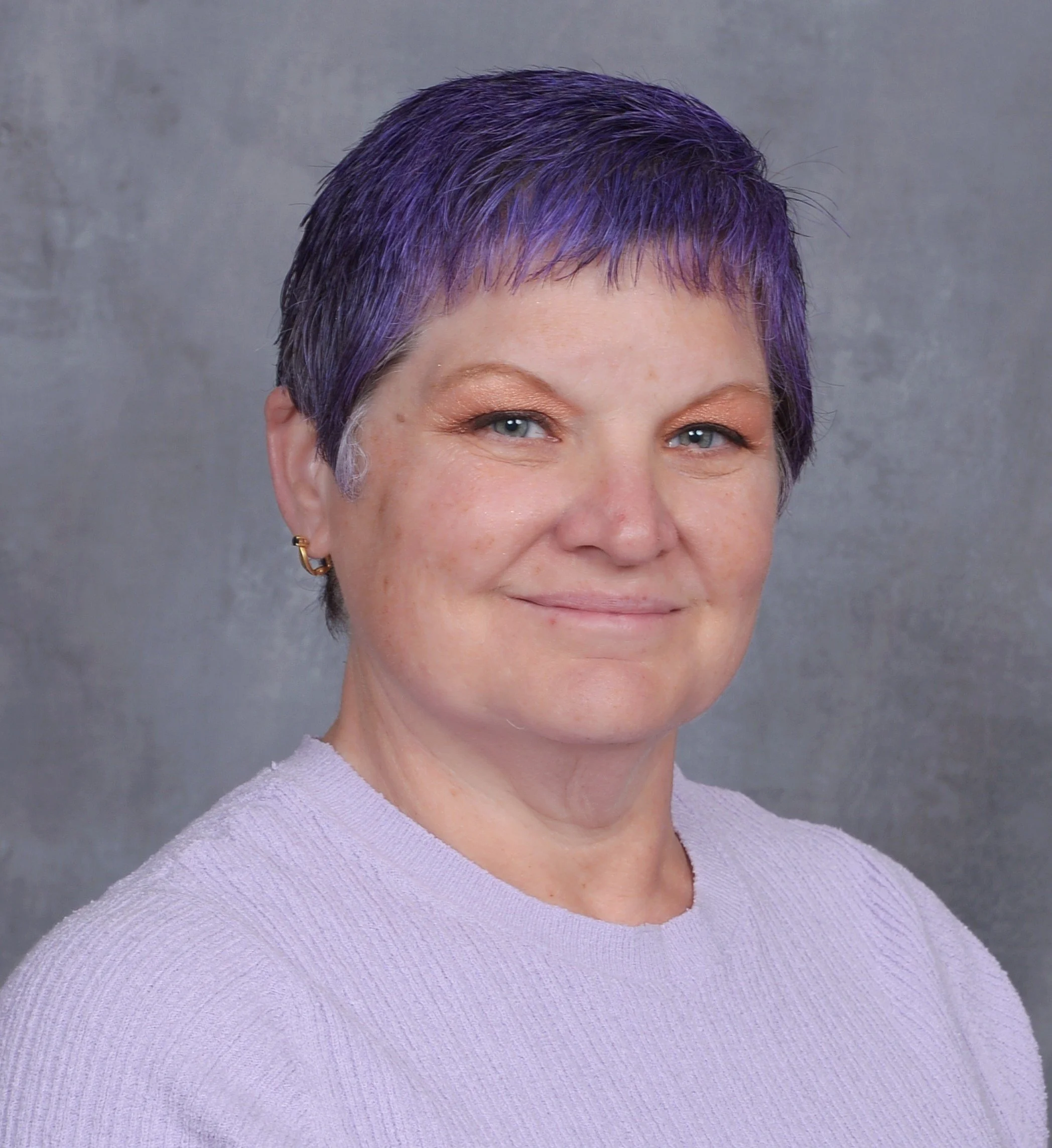 Close-up of a woman's face showing her eyes, nose, and part of her mouth, with short purple hair and wearing gold earrings, dressed in a light purple sweater.