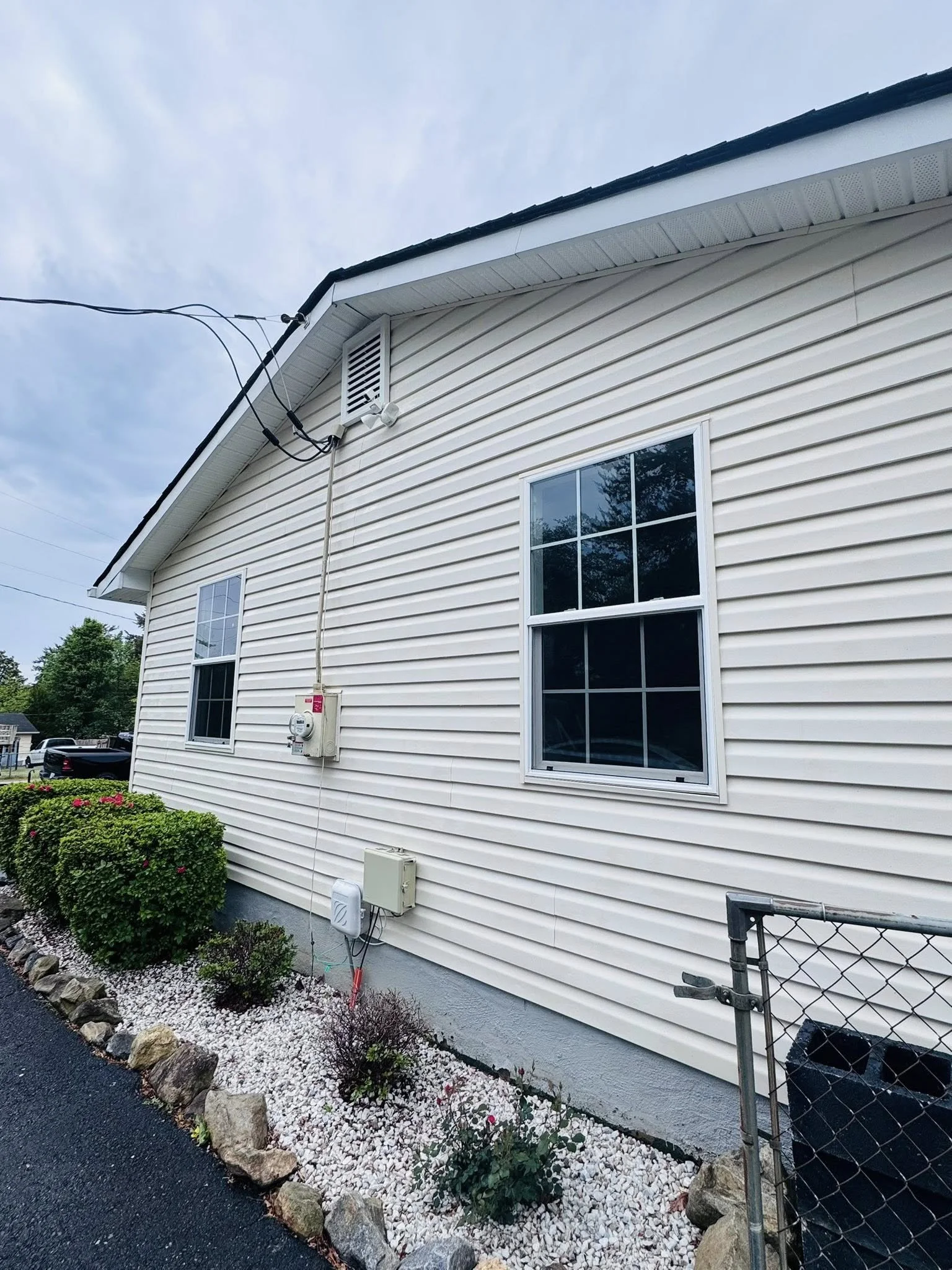 The side of a white house with vinyl siding, two windows, a vent, and outdoor electrical and utility boxes. There are bushes and rocks along the walkway near the house.