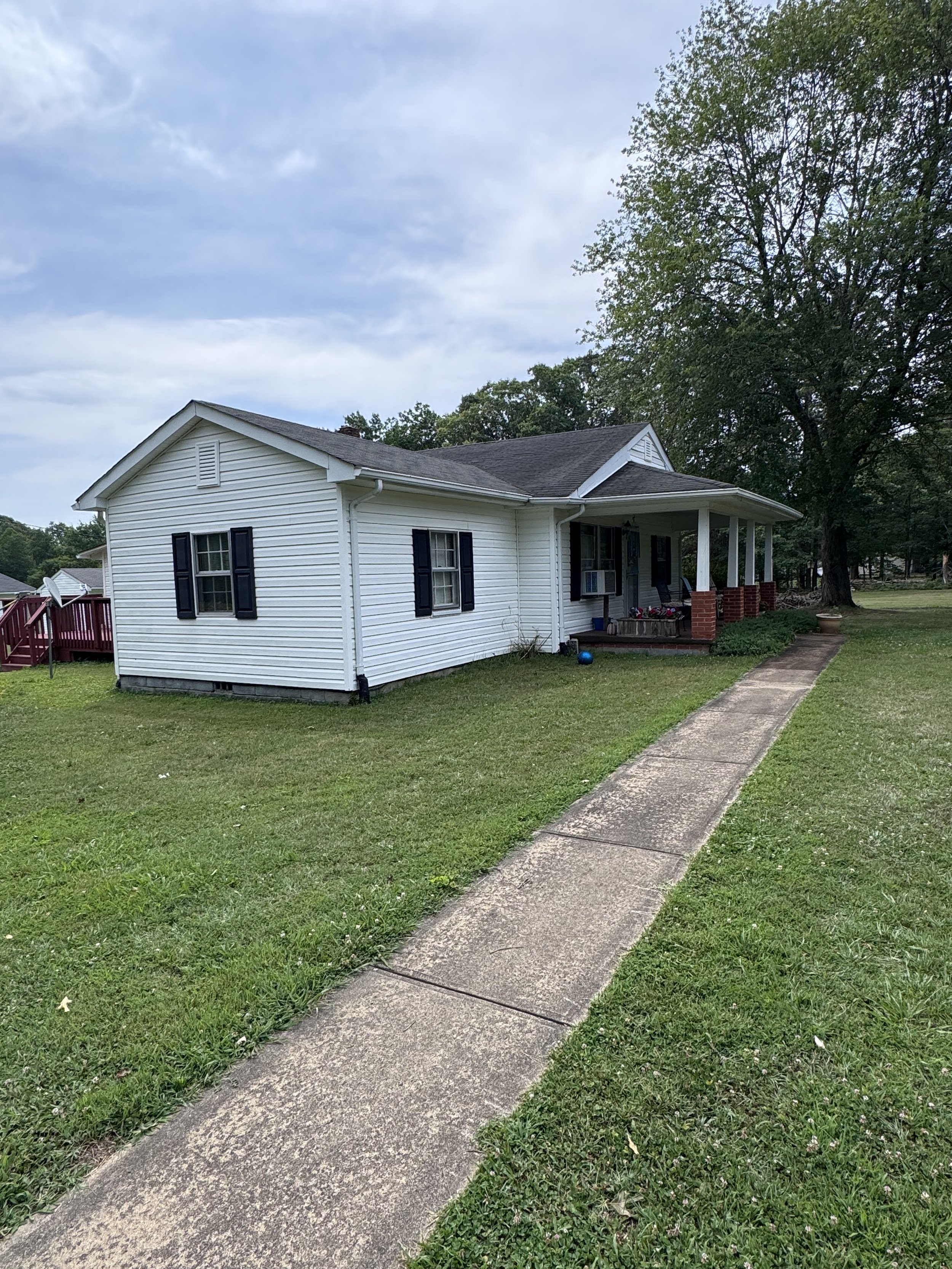 A white single-story house with black shutters and a small covered front porch, surrounded by green grass and large trees. There is a concrete sidewalk leading to the porch, and the sky is cloudy.