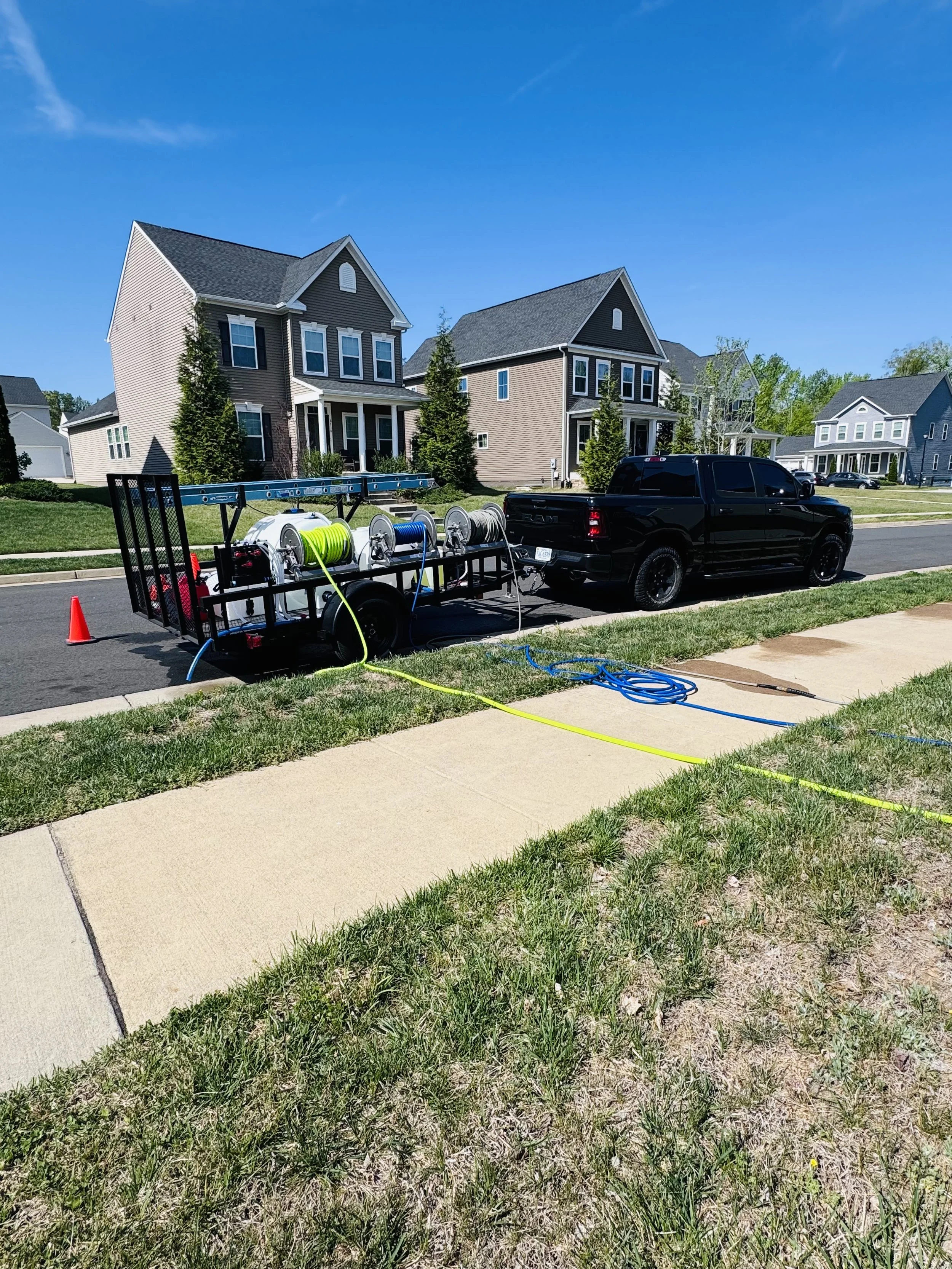 A neighborhood street with several two-story houses, a black pickup truck parked on the street, a trailer with equipment, and green grass in the foreground on a clear, sunny day.