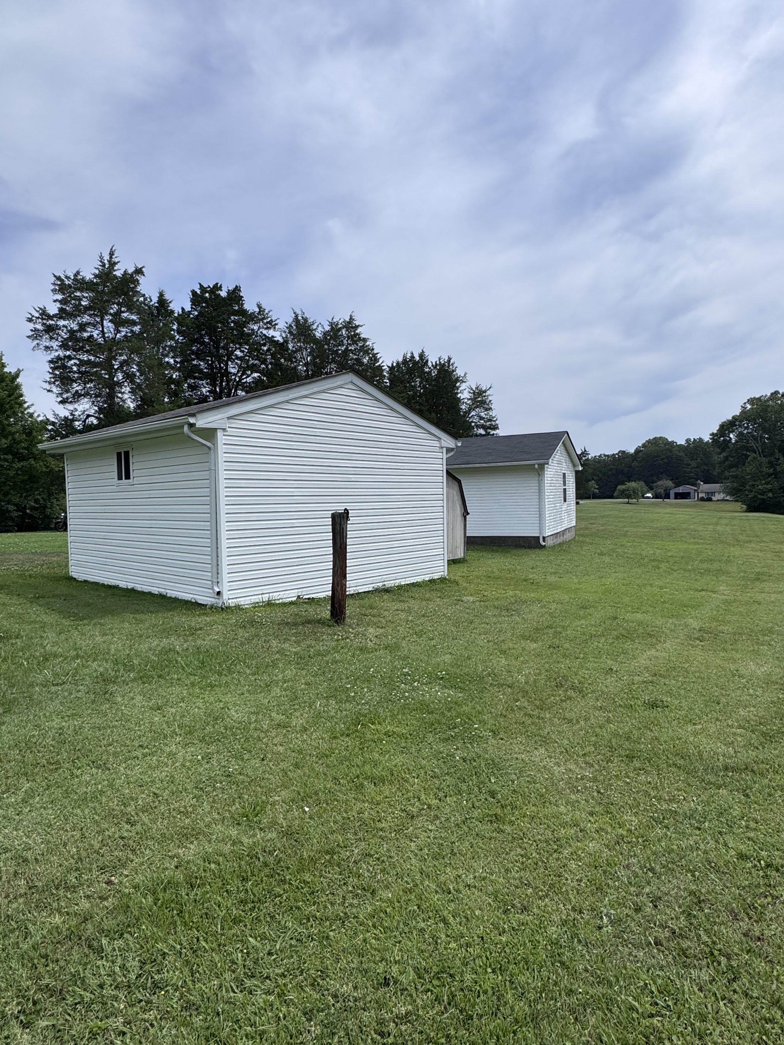 A white house with vinyl siding in a grassy field under a cloudy sky. There are trees in the background and a smaller structure attached to the house.