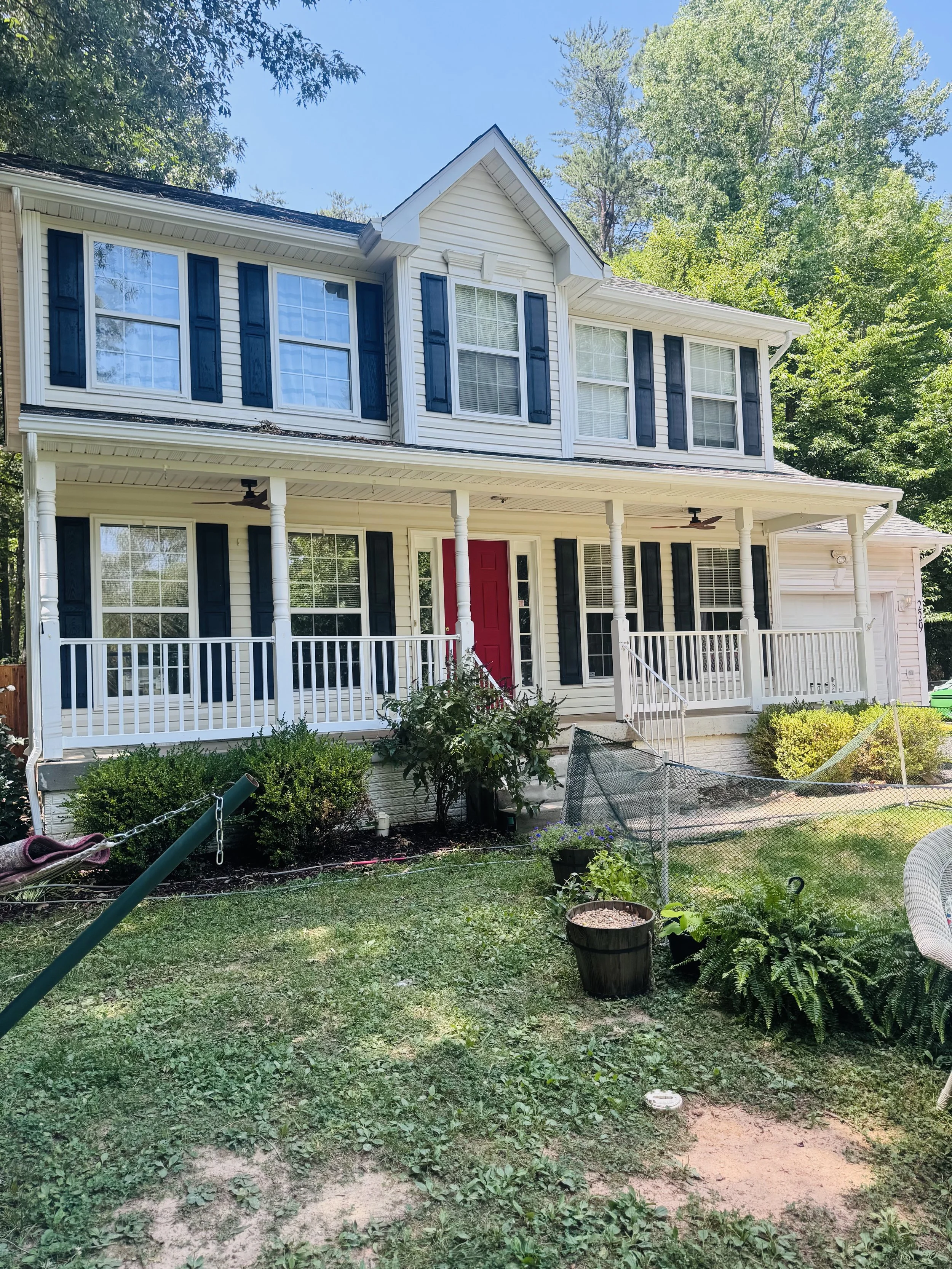 A two-story house with white siding, black shutters, and a red front door, surrounded by a garden with various plants and trees, a white railing on the porch, and a clear blue sky in the background.