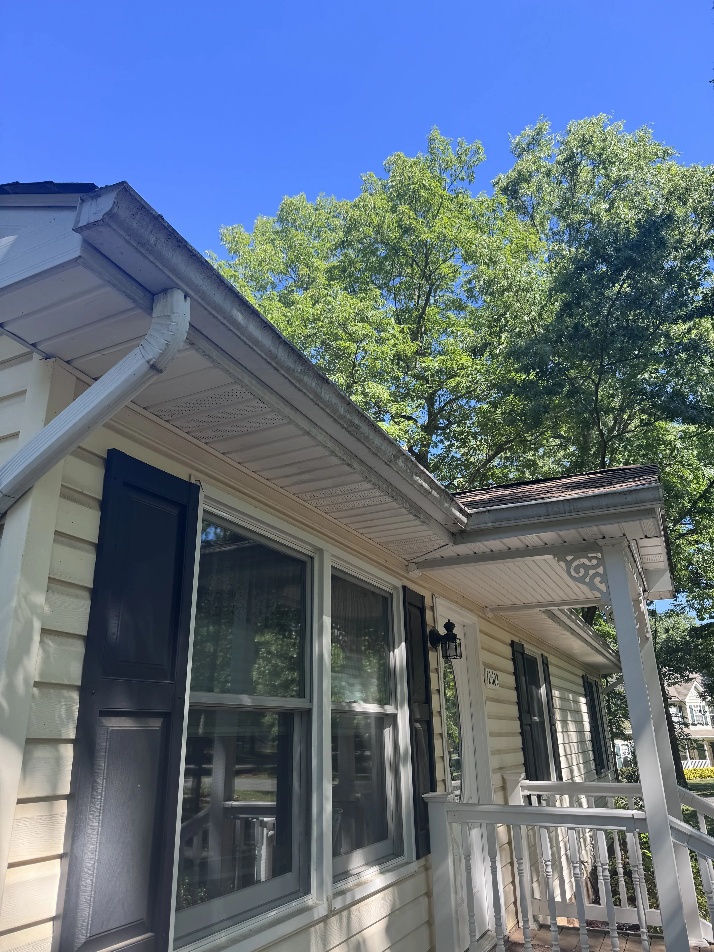 View of a house exterior with beige siding, black shutters, and three large windows, a white porch railing, and a blue sky with green trees in the background.