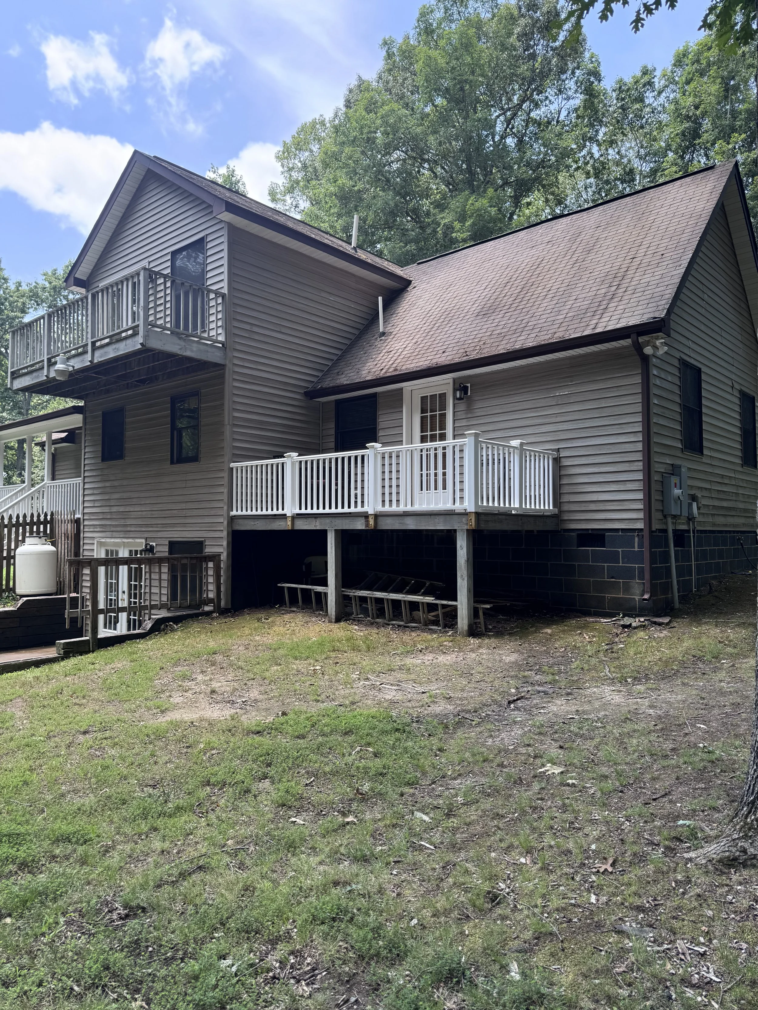 A two-story house with gray siding, two decks, one on each level, surrounded by green trees and a lawn with patches of dirt and grass.