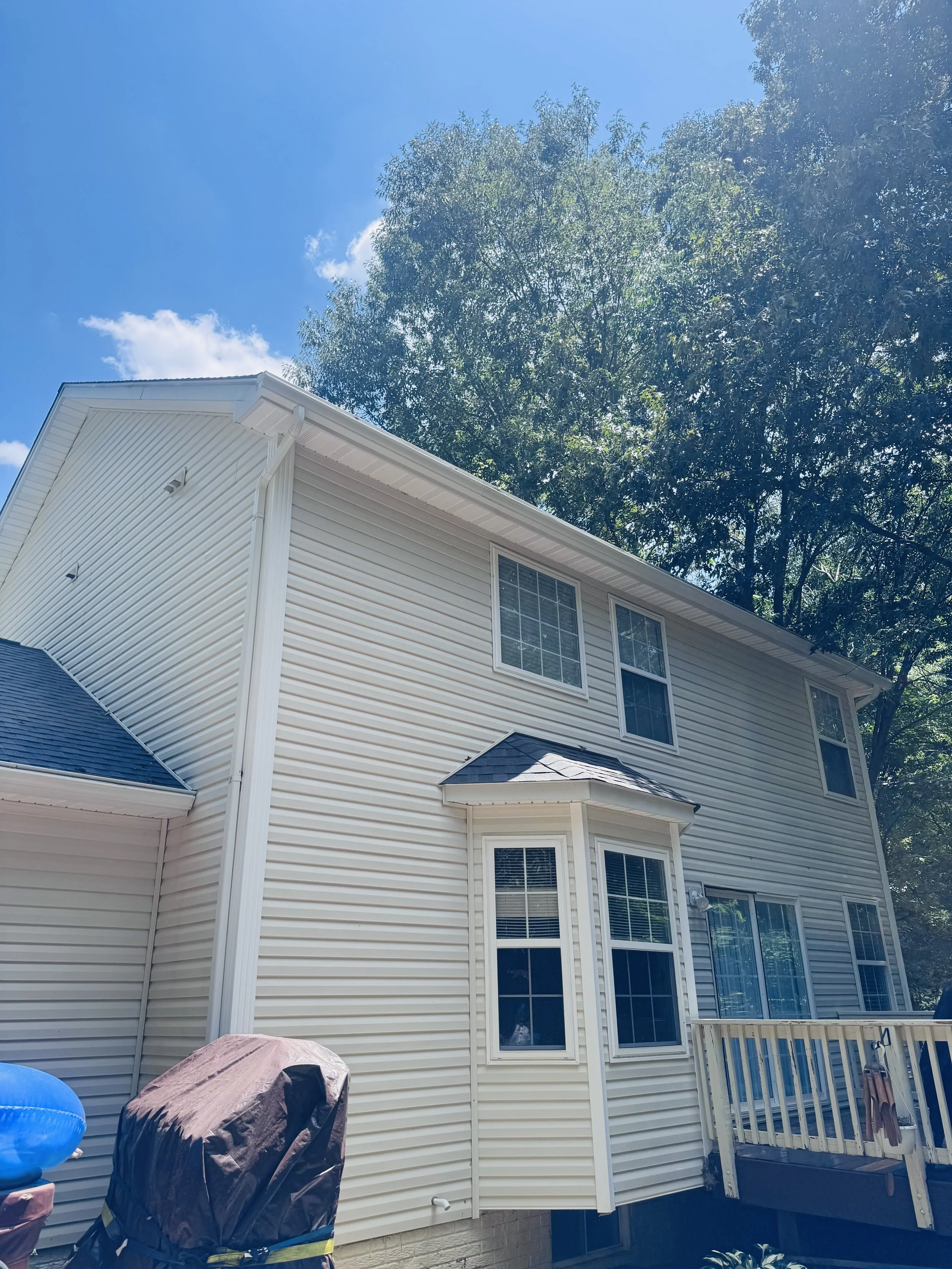A two-story house with beige vinyl siding, several windows, and a small porch with a white railing. There are trees in the background, and the sky is clear with a few clouds. In the backyard, there is a covered grill and a blue object.