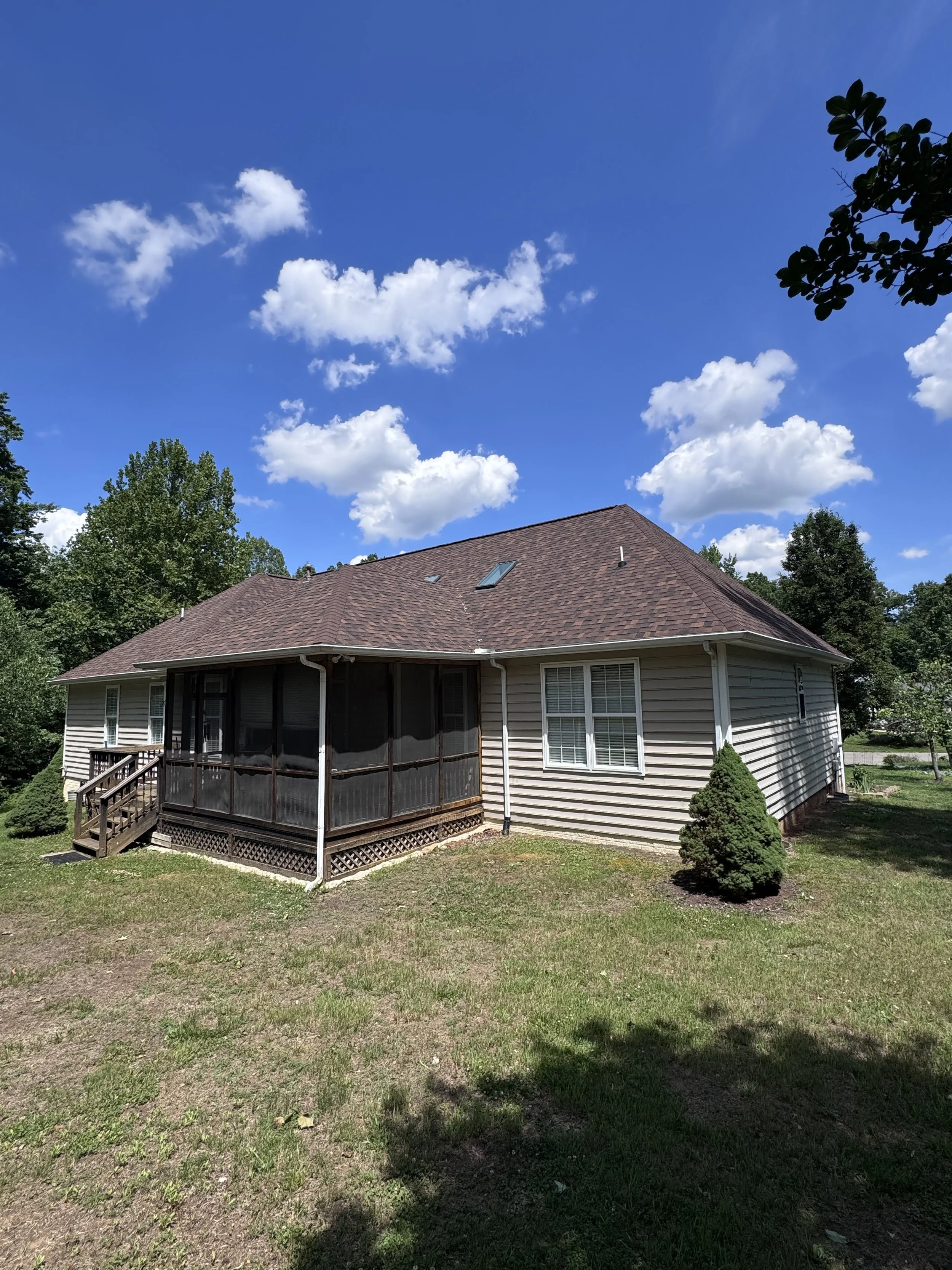A house with beige siding, a brown shingled roof with skylights, and a screened porch in a lush green yard with trees and cloudy blue sky.