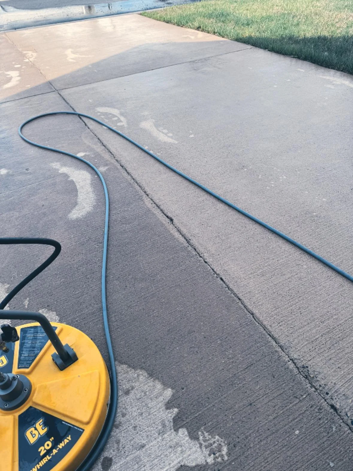 A wet concrete driveway with a yellow wet saw and a blue hose lying on it, with some water spray and footprint marks, and a grassy lawn at the edge.