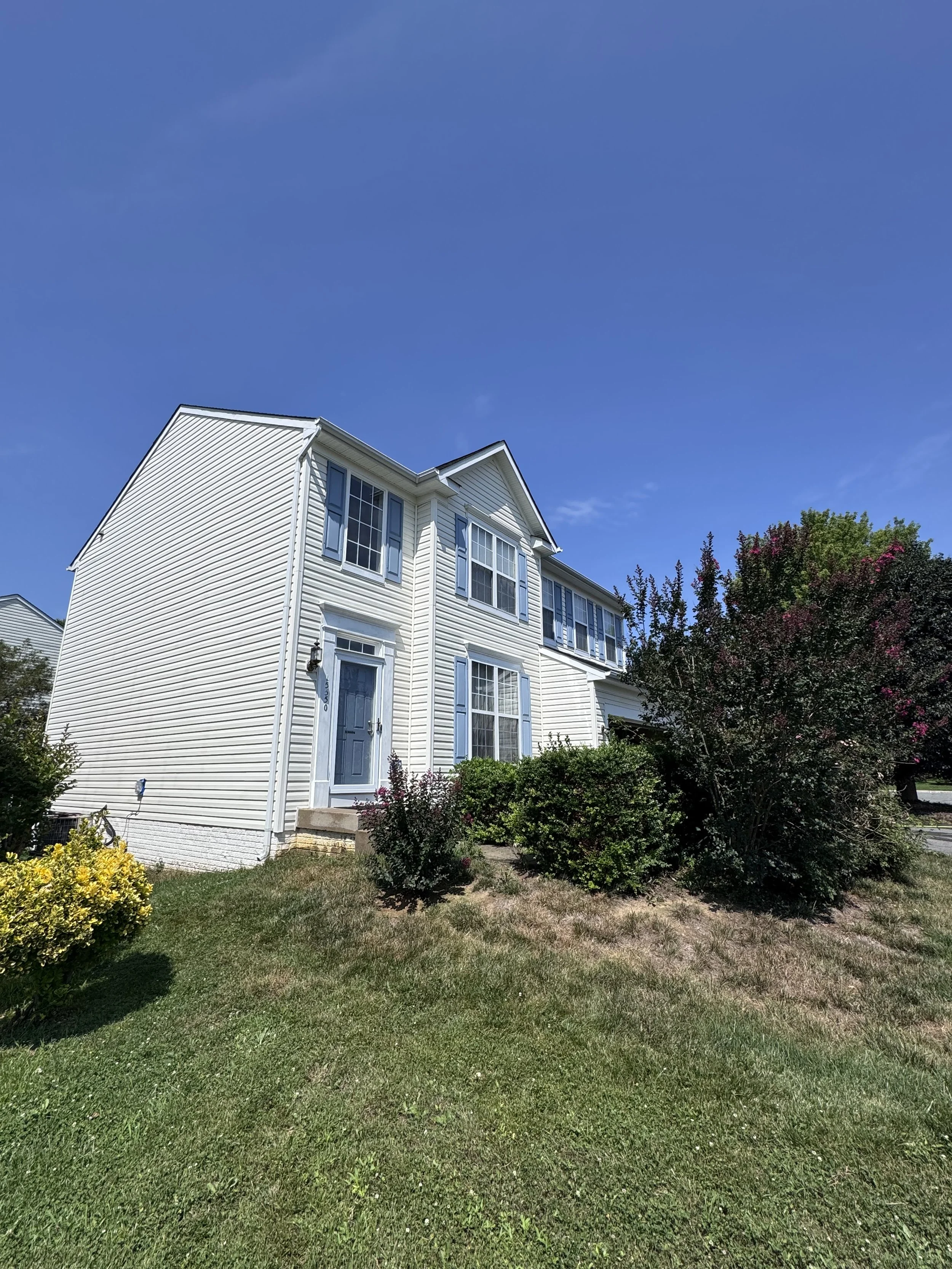 A white two-story house with blue shutters and front yard garden, trees, and a clear blue sky.