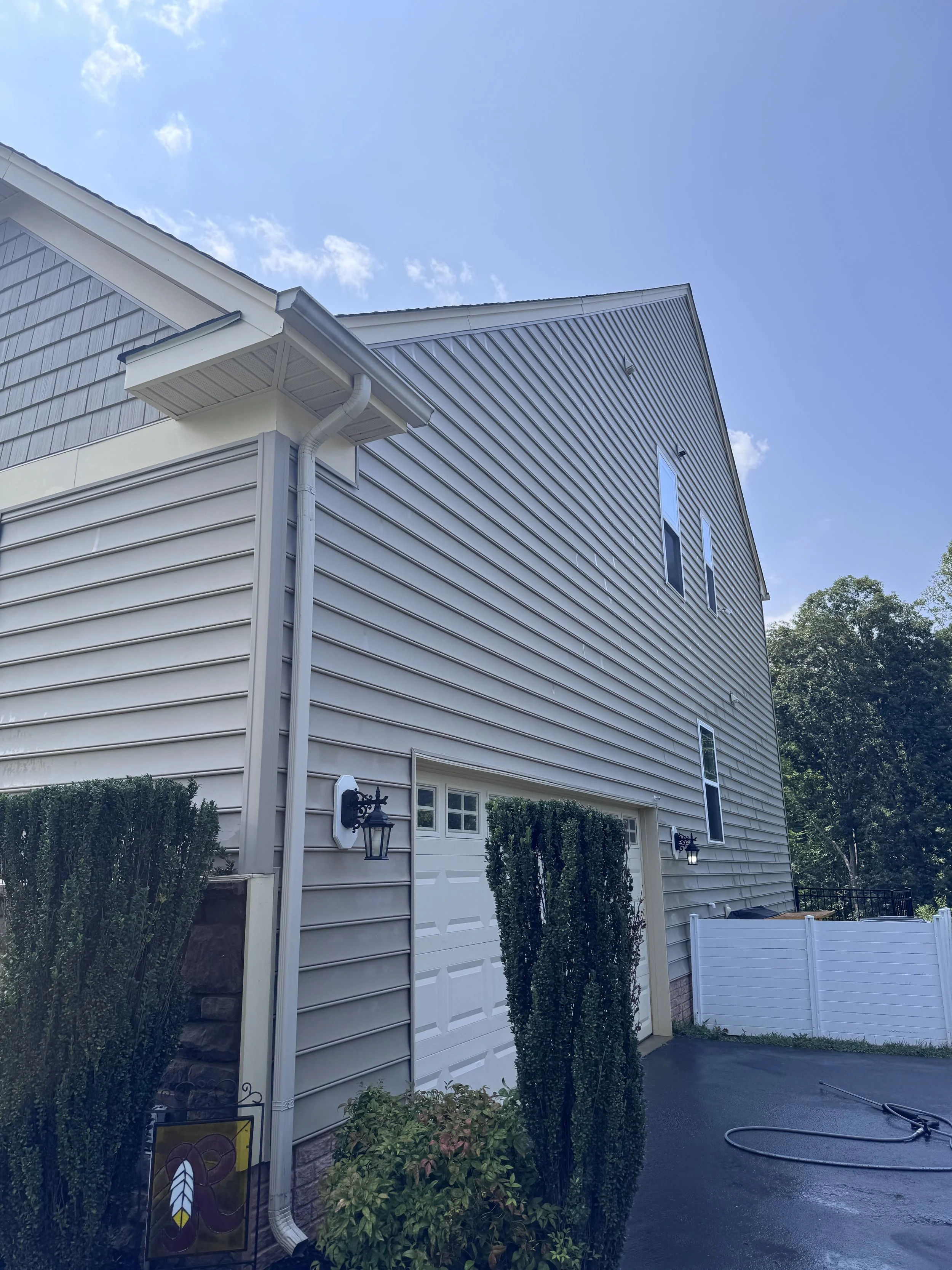 Front of a light-colored house with a garage door, two black wall-mounted lanterns, and a garden with bushes and a small sign. The house has vertical siding and a sloped roof with gutters. Clear sky and trees in the background.