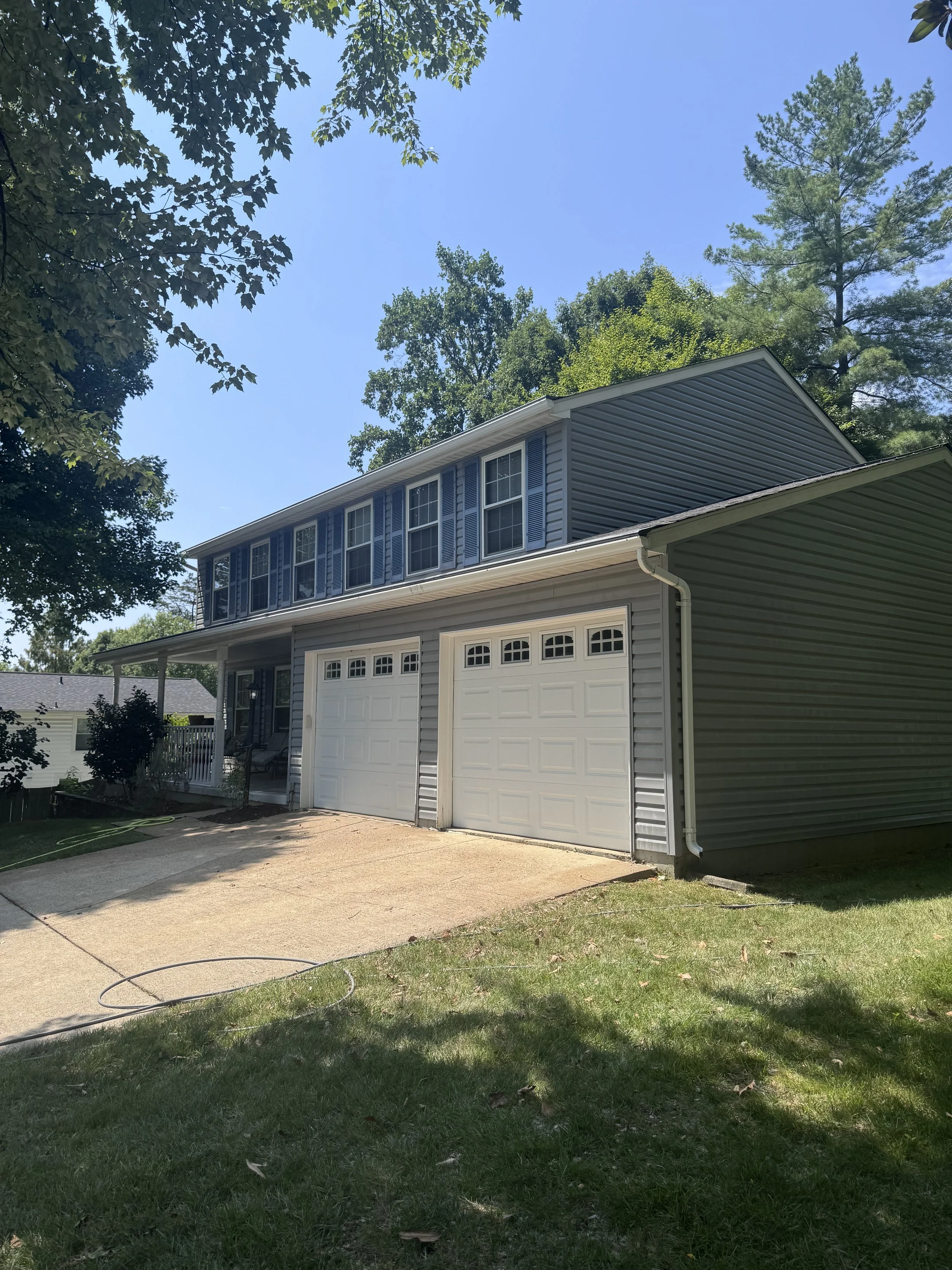A two-story house with gray siding and white garage doors, surrounded by trees under a clear blue sky.