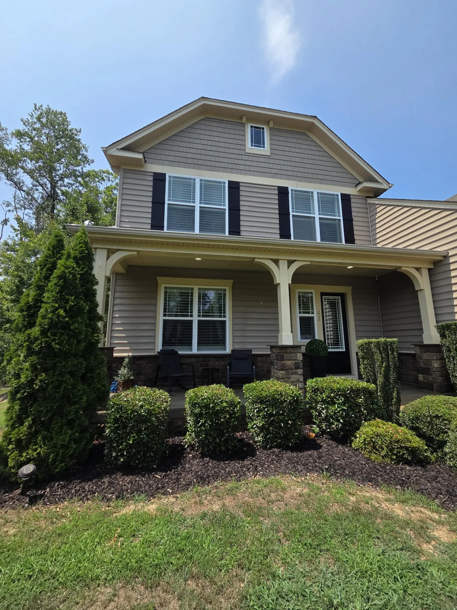 Front view of a two-story house with beige siding, white trim, black shutters, large windows, a covered porch with columns, surrounded by greenery and bushes, with trees and a blue sky in the background.