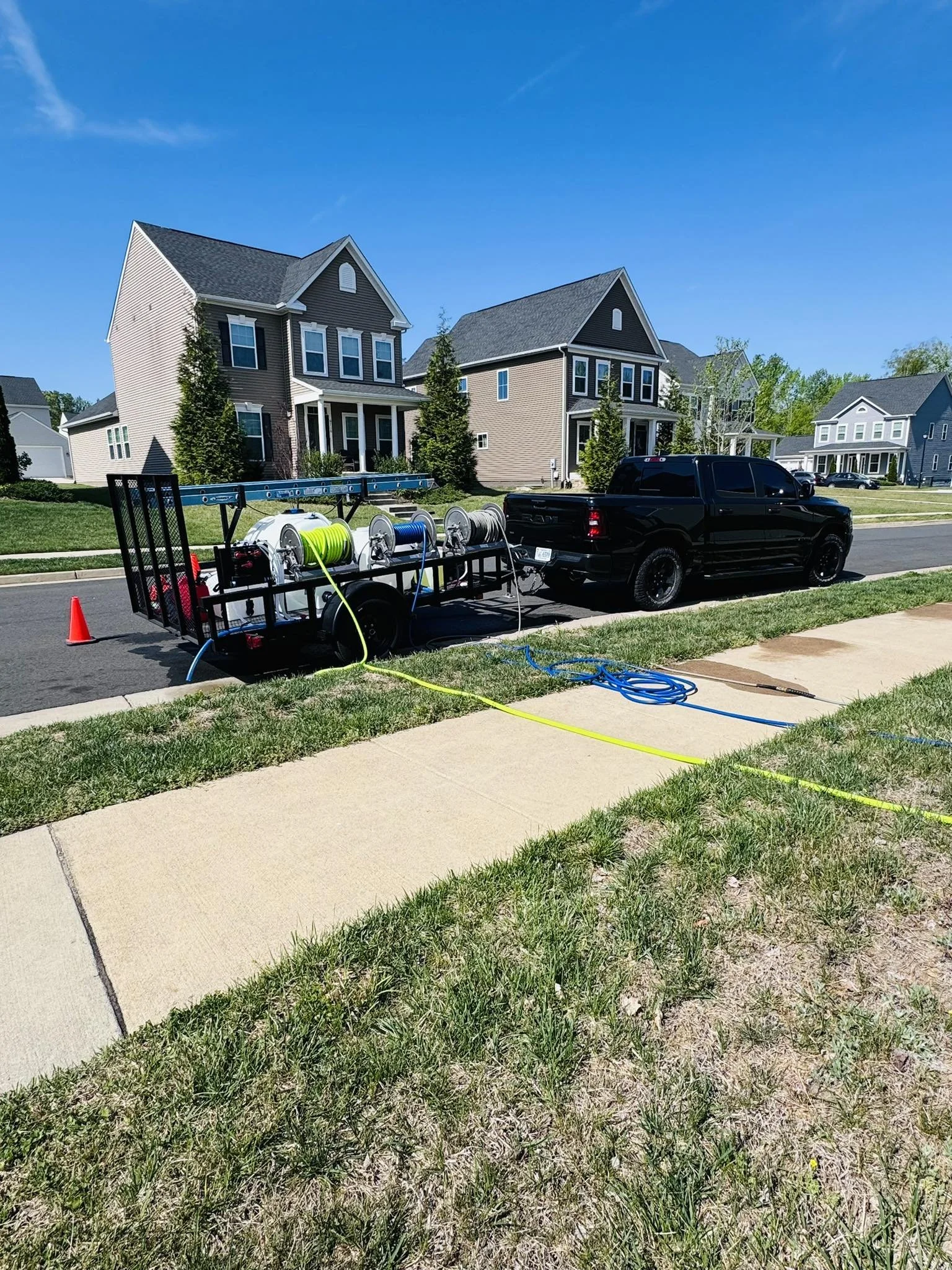 A residential street on a sunny day with houses, a black pickup truck, and a trailer carrying hoses and equipment, with green grass and sidewalk in the foreground.
