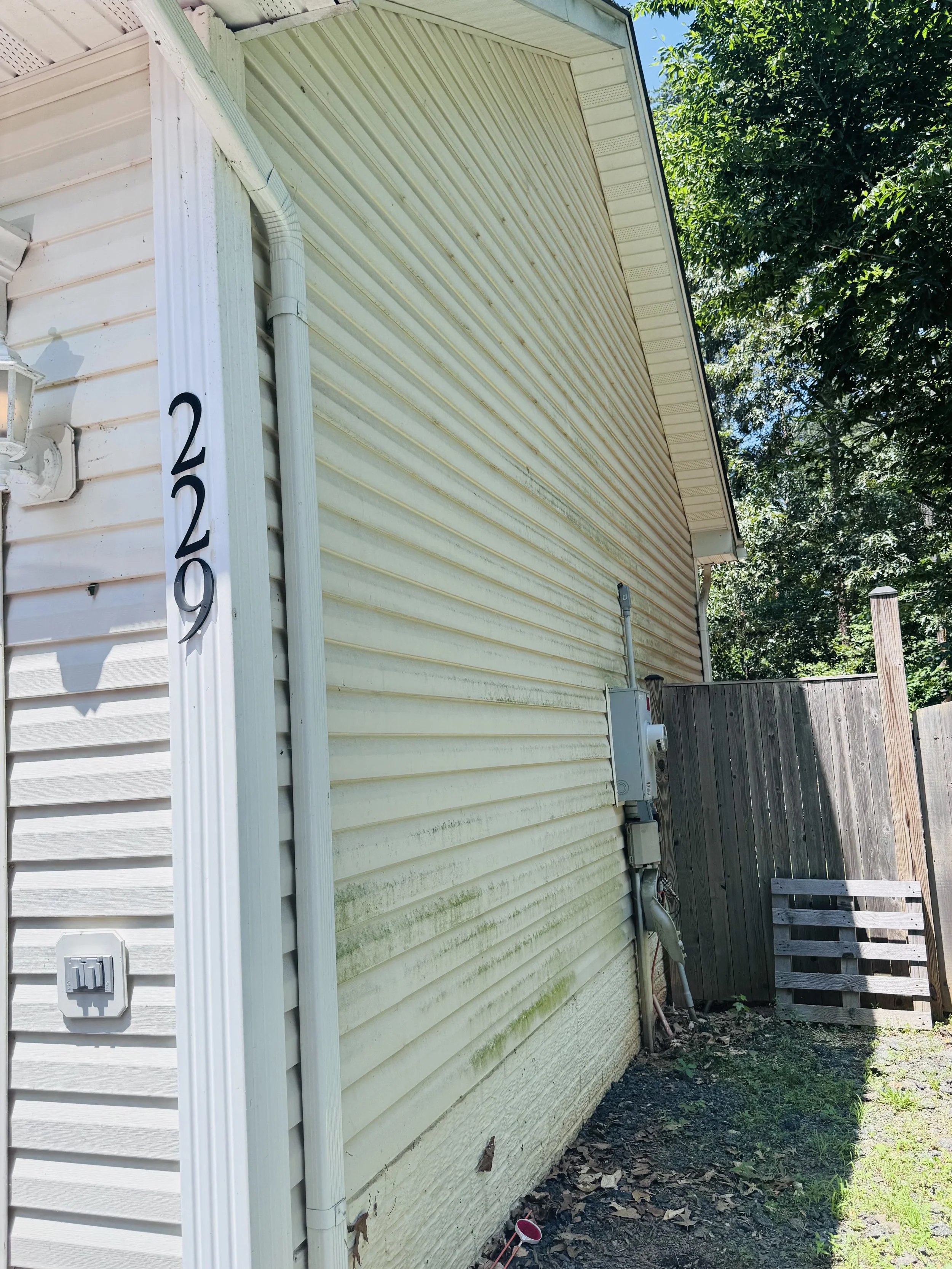 The side of a house with beige vinyl siding, a white rain gutter, electrical meter, and an outdoor fence with green plants and trees nearby.