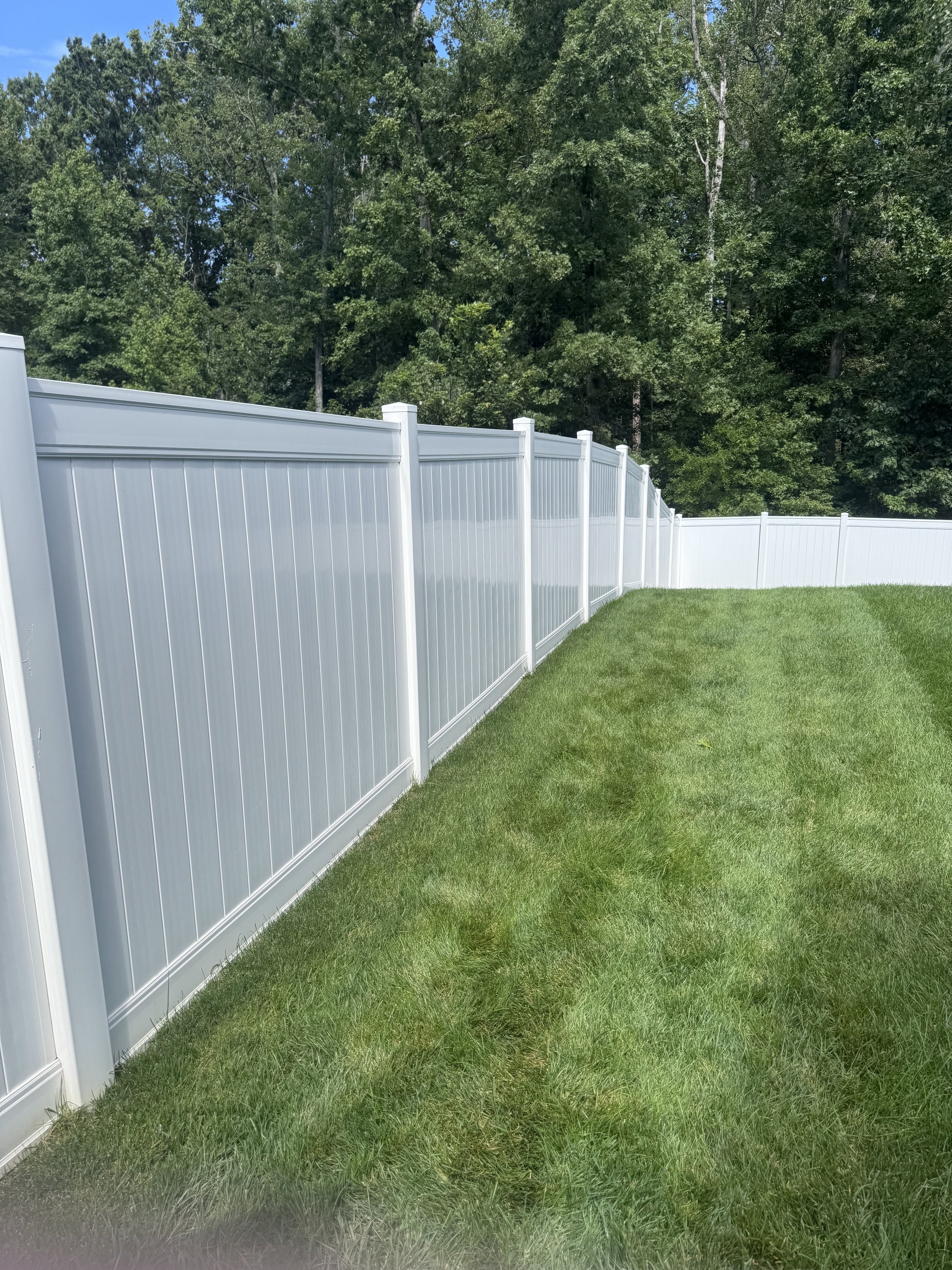 A white vinyl privacy fence runs alongside a well-manicured green lawn with a dense row of trees in the background under a bright, partly cloudy sky.