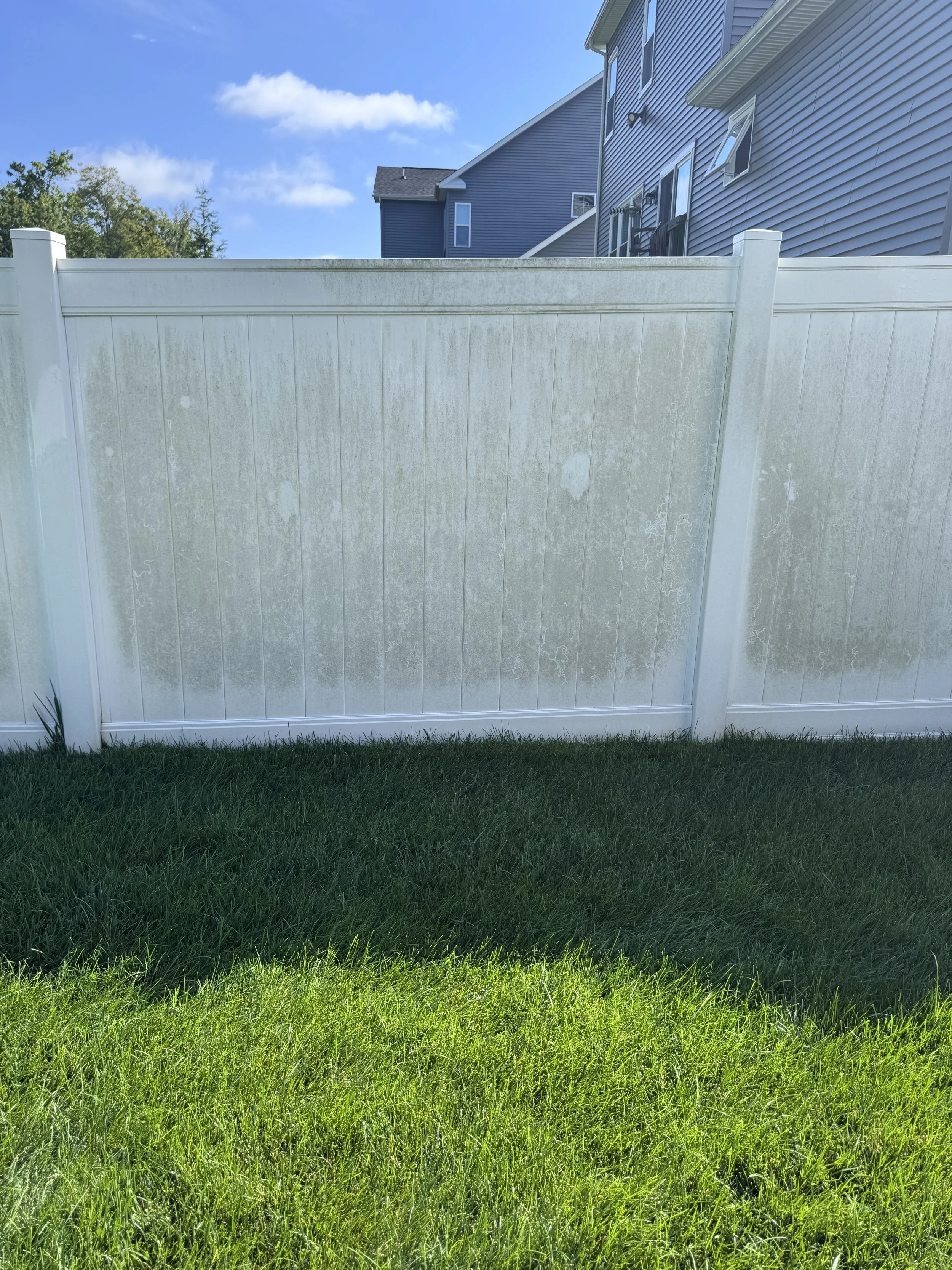 A white privacy fence with some dirt and moss on top, in front of a grassy yard. There are blue houses with windows and balconies in the background, and a blue sky with clouds overhead.