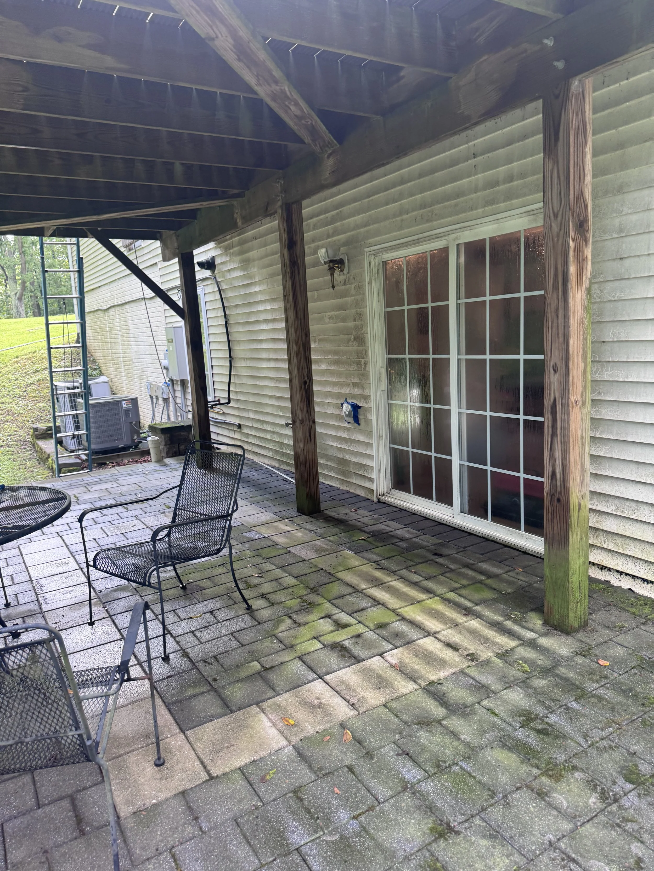 Backyard patio with black metal furniture, pavers with moss and leaves, and part of the house wall with sliding glass door and outdoor light fixture.