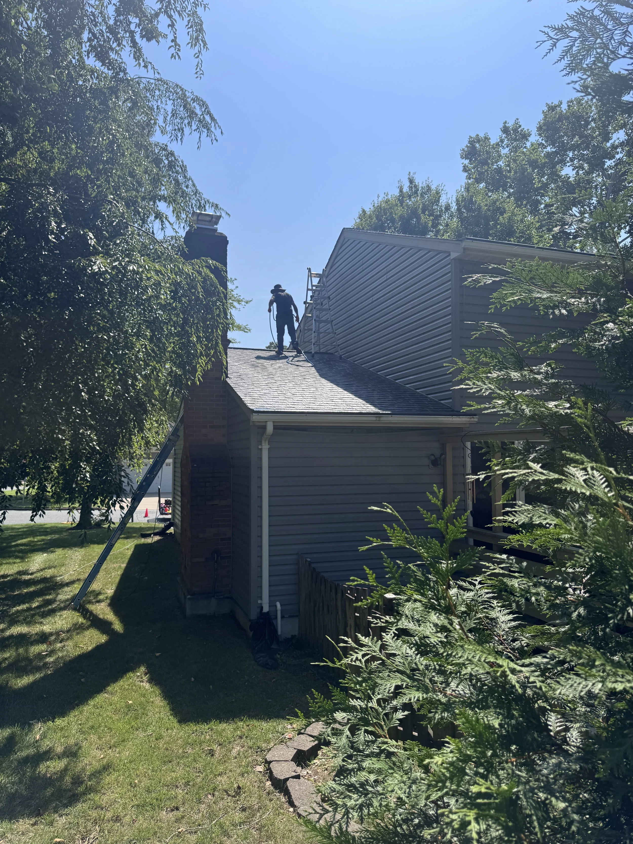 A person working on the roof of a house during daytime, standing near a ladder with trees and a blue sky in the background.