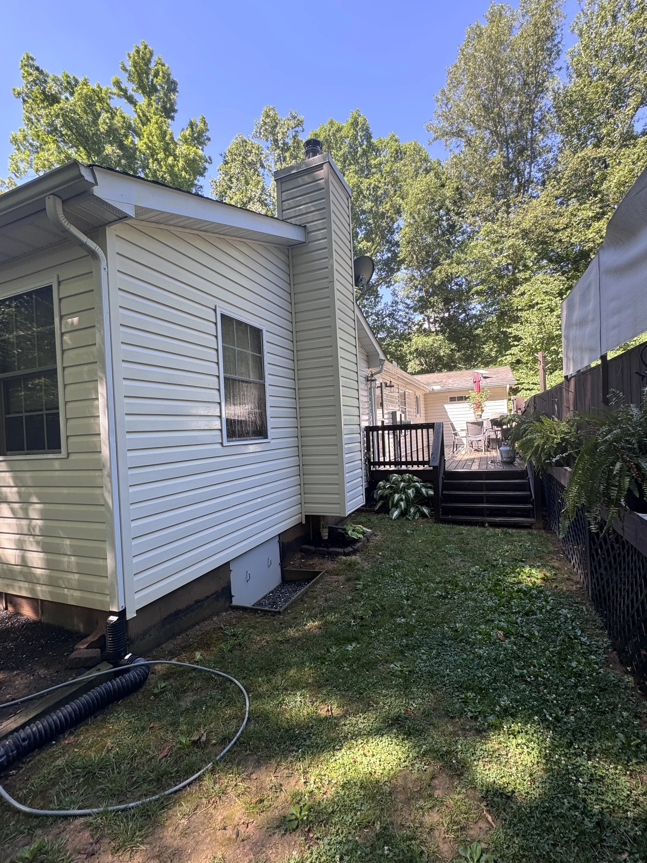Backyard with a house featuring white siding, a deck with outdoor furniture and an umbrella, trees, and a bright blue sky.